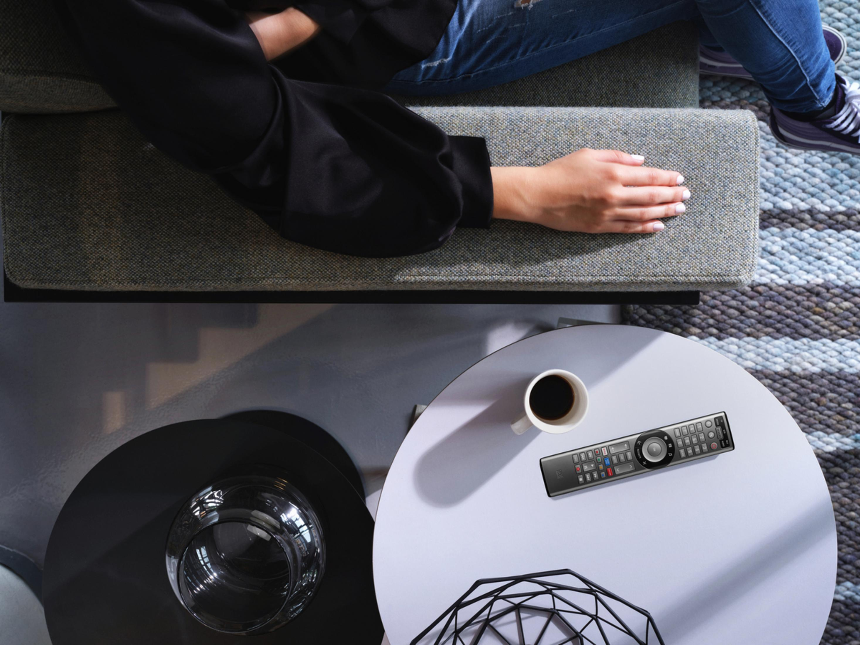 Eine Person sitzt auf einem Sofa, eine Hand auf dem Kissen, Fernbedienung und Kaffee auf dem Tisch.