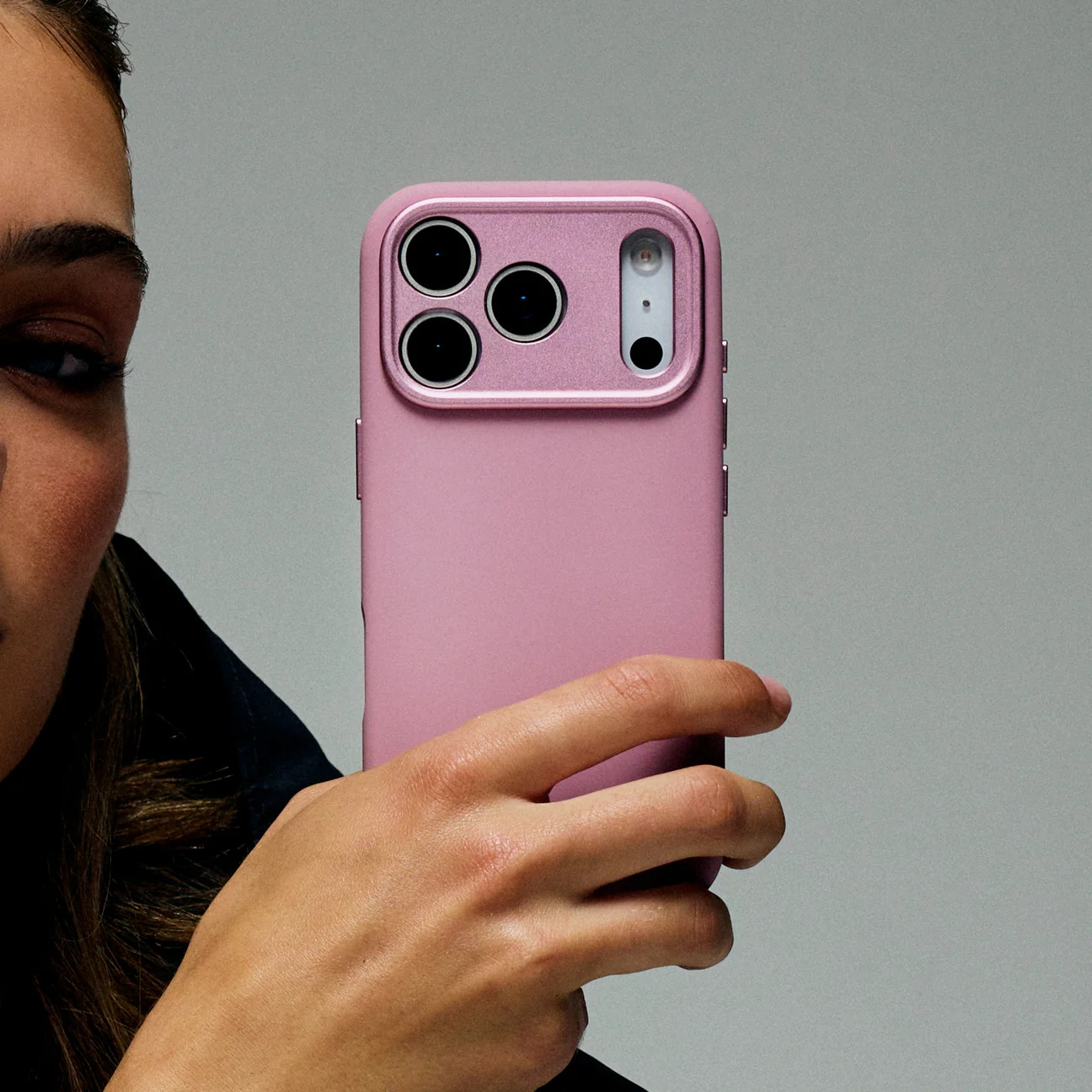 Eine Hand hält ein rosafarbenes Smartphone mit drei Kameras vor einem grauen Hintergrund.