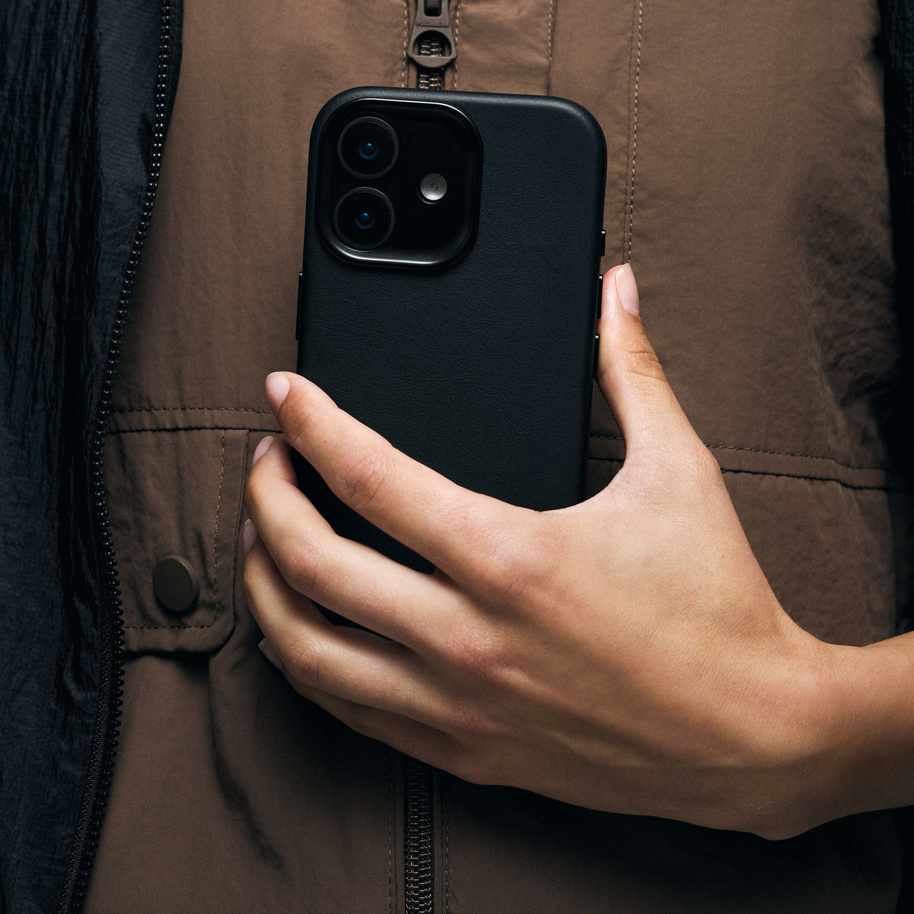 Una mano tiene uno smartphone nero contro una giacca marrone con zip.