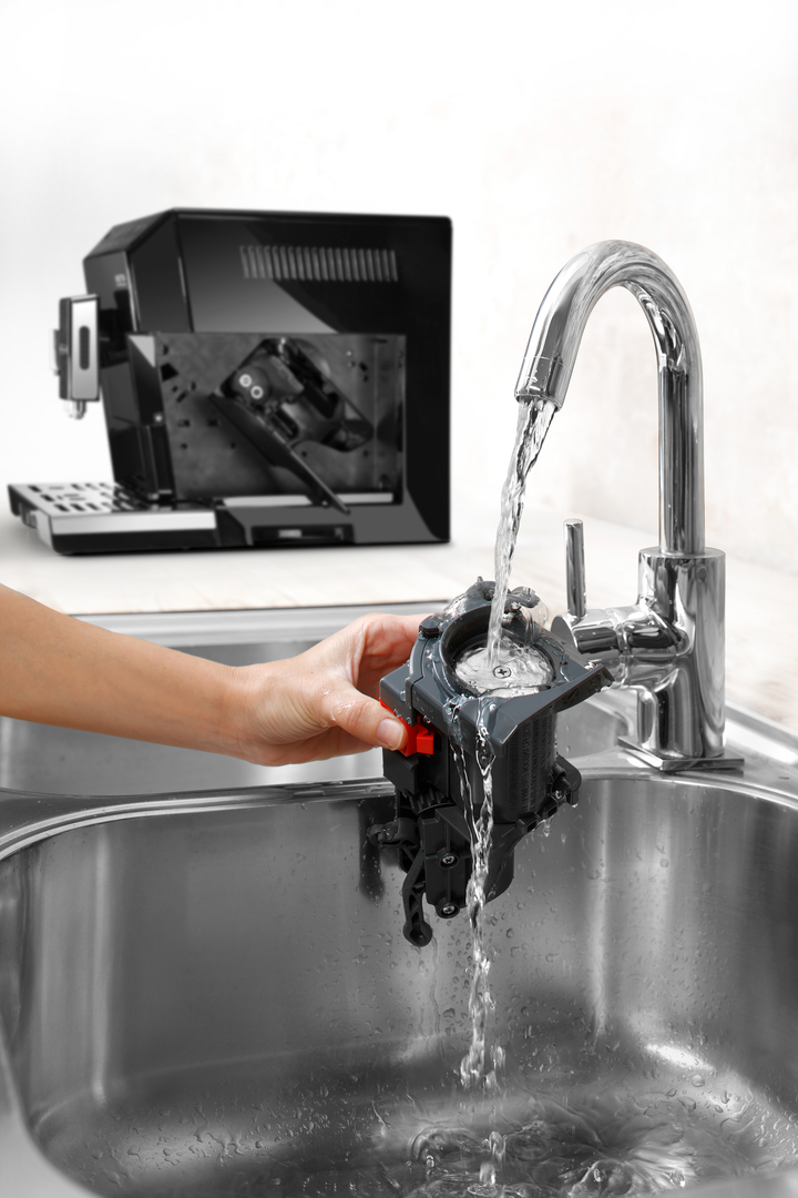 Eine Hand reinigt eine Kaffeemaschinenkomponente unter fließendem Wasser in einem Spülbecken.