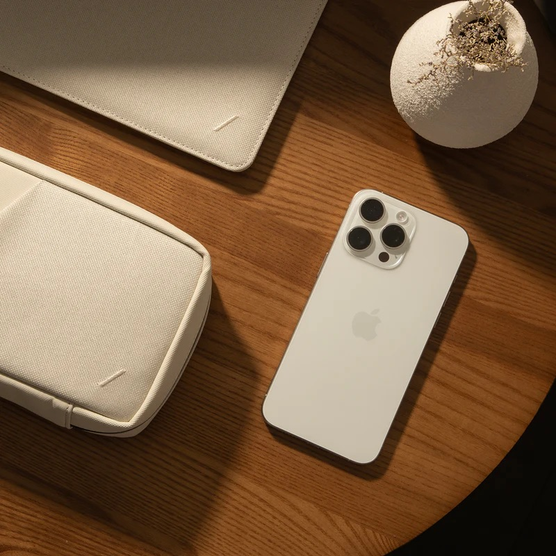 Un iPhone bianco giace su un tavolo di legno accanto a una pochette beige e un vaso.