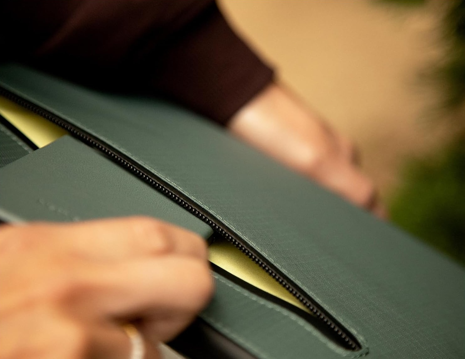 Mani aprono una pochette verde con cerniera, fodera gialla visibile.
