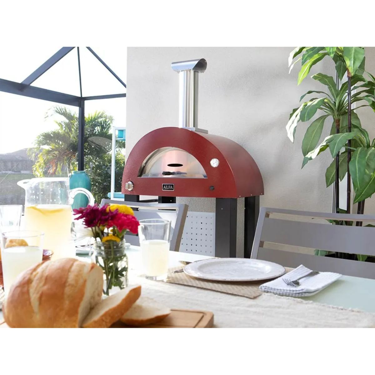 Ein roter Pizzaofen steht neben einem Esstisch mit Brot, Getränken und Blumen.