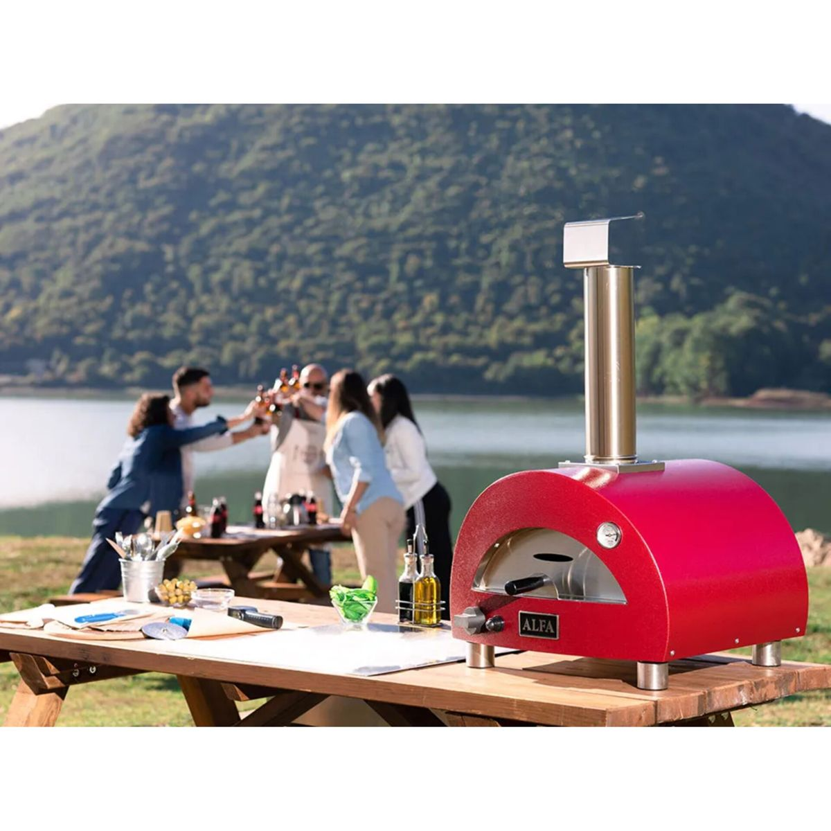 Rote Pizzaofen auf einem Holztisch im Freien, Leute stoßen an, See und Hügel im Hintergrund.
