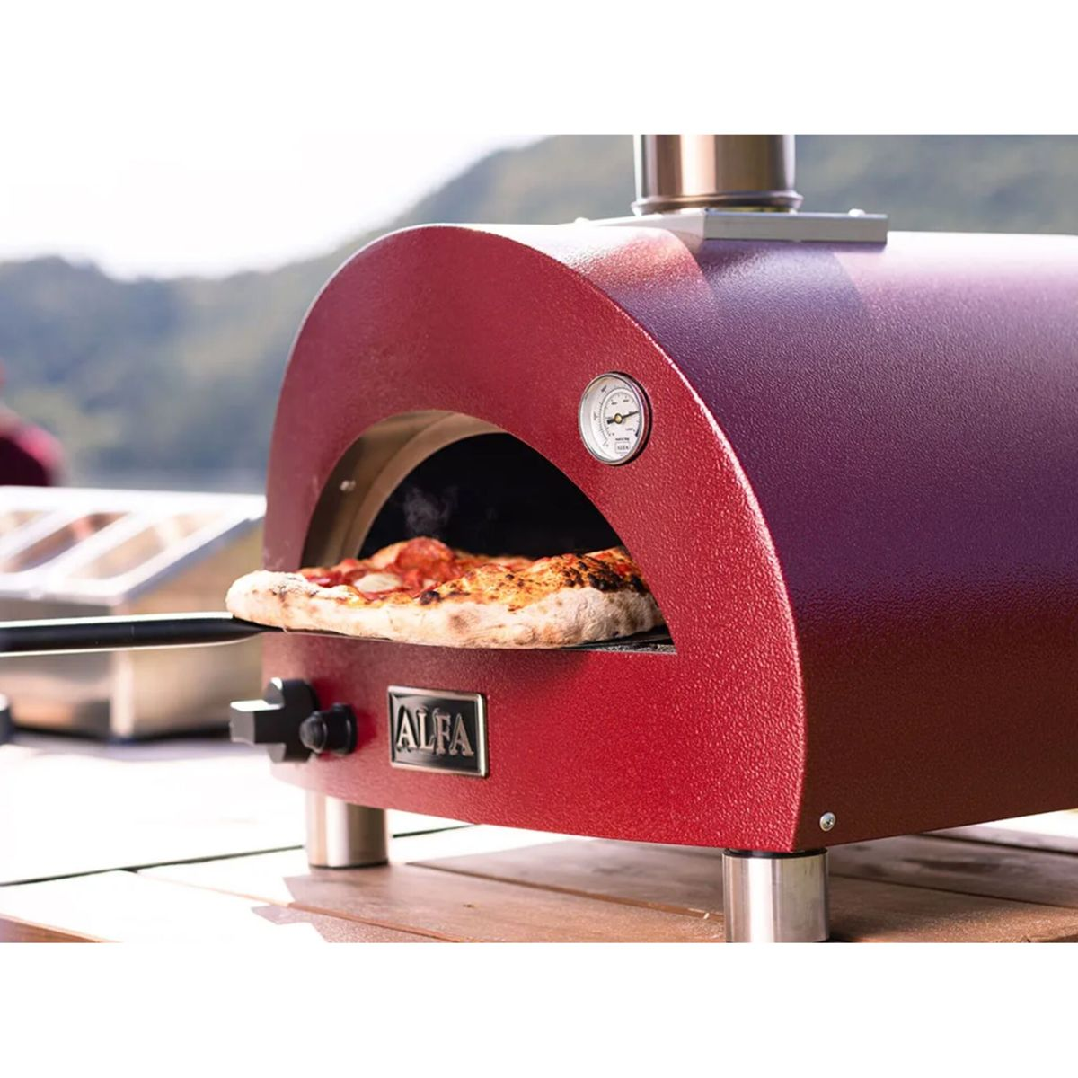 Ein roter Pizzaofen mit einem Gargericht wird herausgezogen. Berge im Hintergrund.