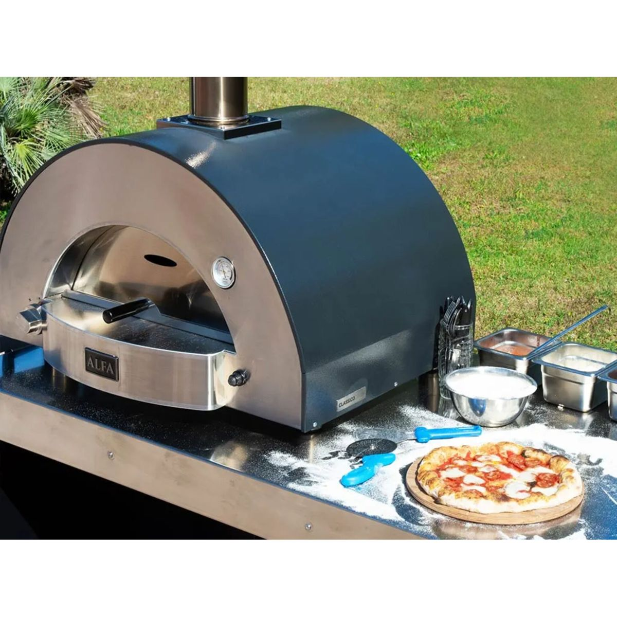 Ein Pizzaofen im Freien mit einer fertigen Pizza auf der Arbeitsplatte. Grüne Grasfläche im Hintergrund.