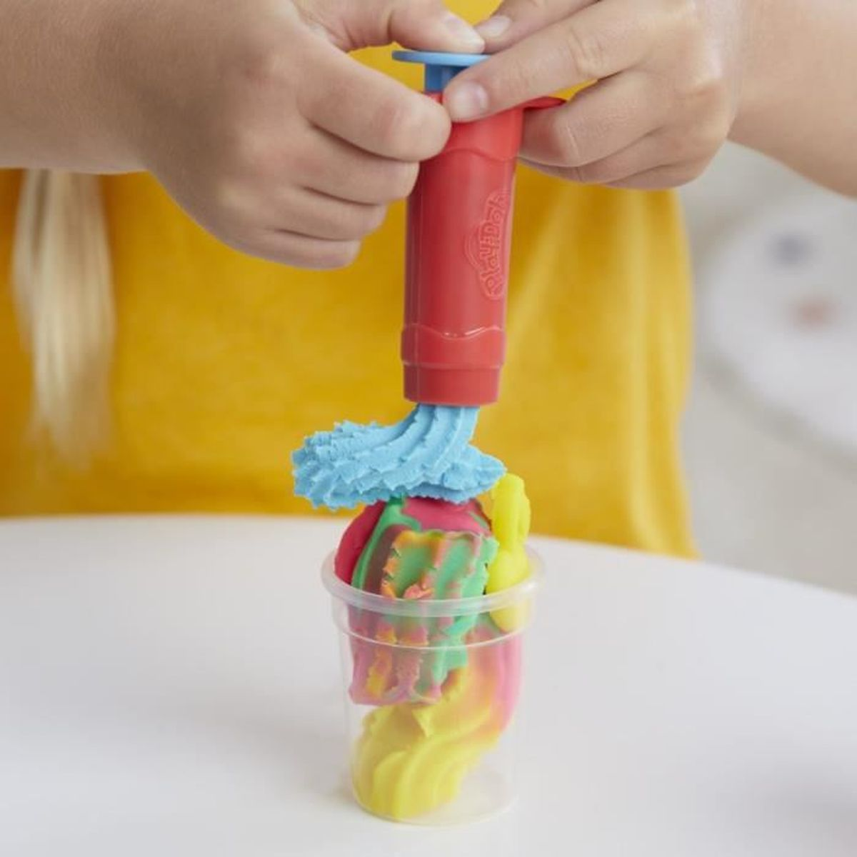 Manos usan un extruidor rojo de Play-Doh para meter masa colorida en un vaso transparente.