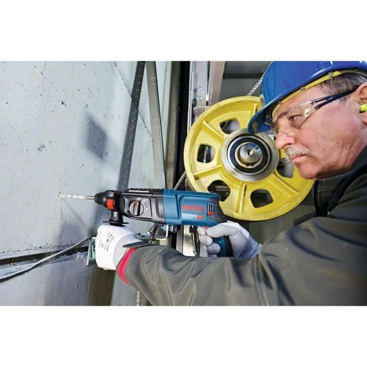 Man with drill bores hole in wall, yellow wheel in background.