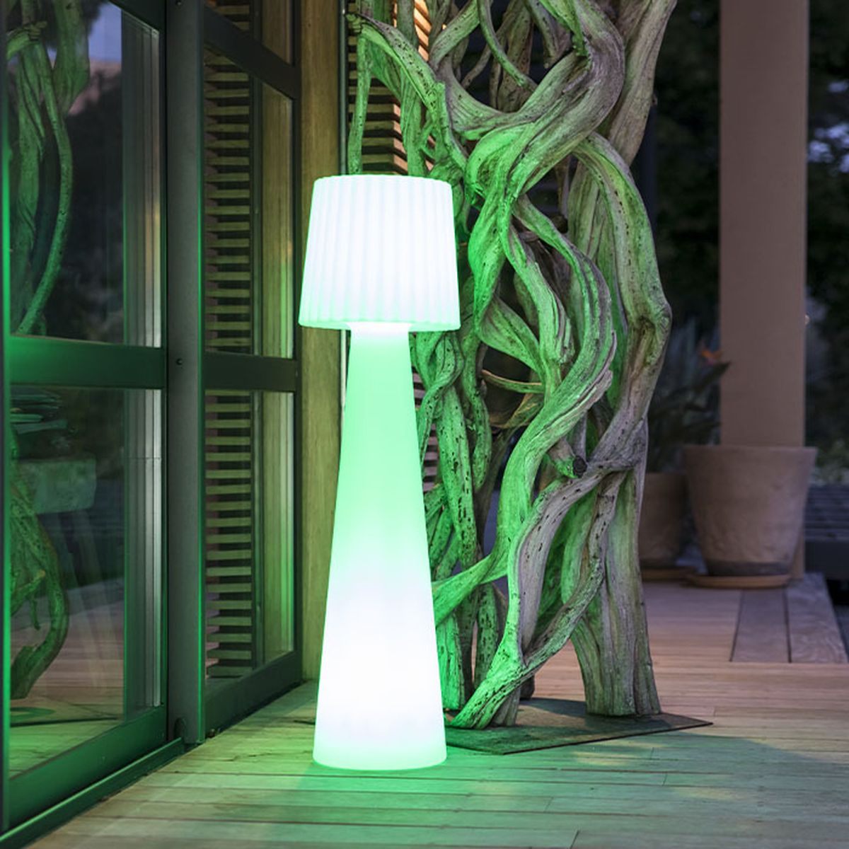Una lampada alta a forma di cono con paralume bianco plissettato emette luce verde.