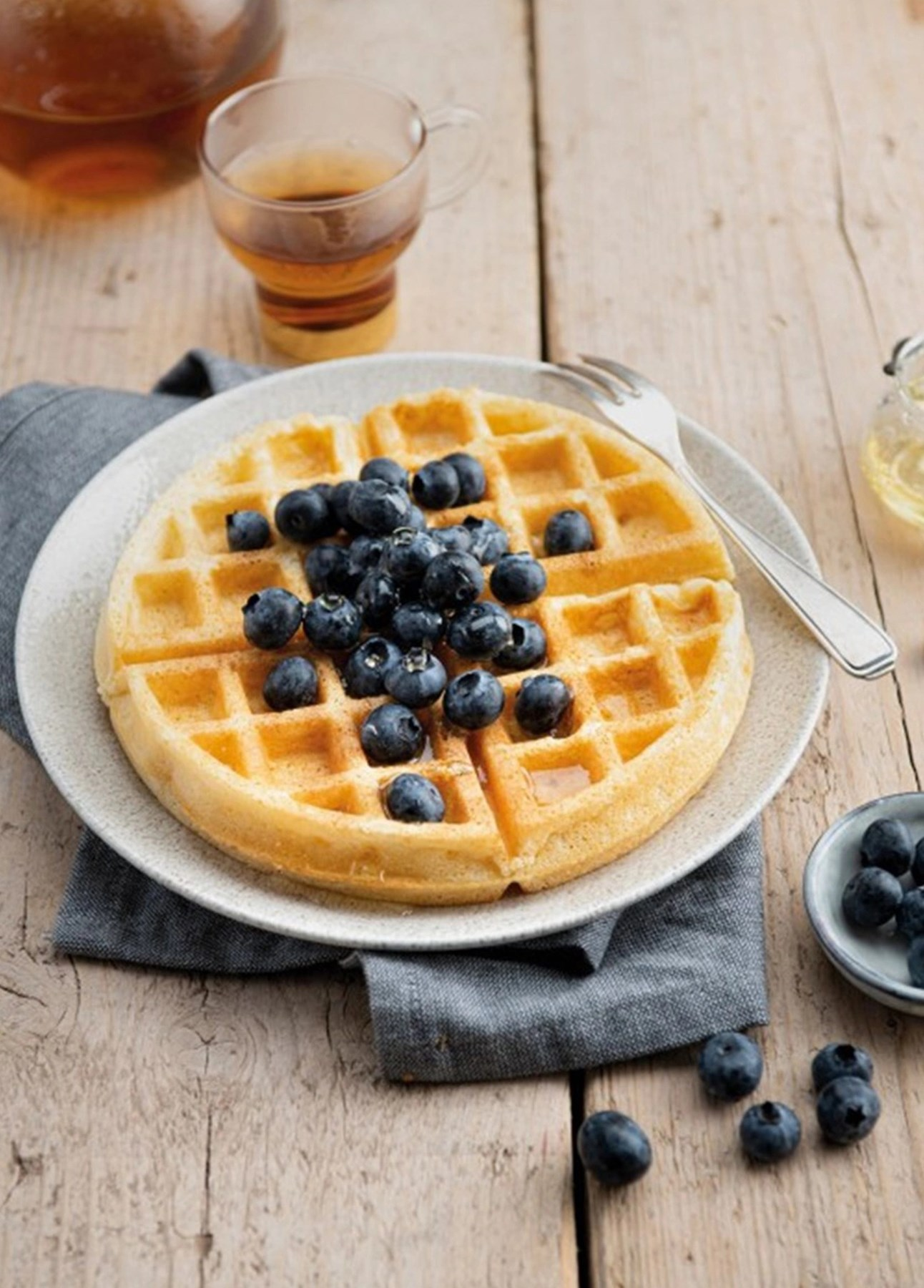 Üzeri yaban mersiniyle kaplı yuvarlak bir waffle, çay ve balın olduğu tabakta.