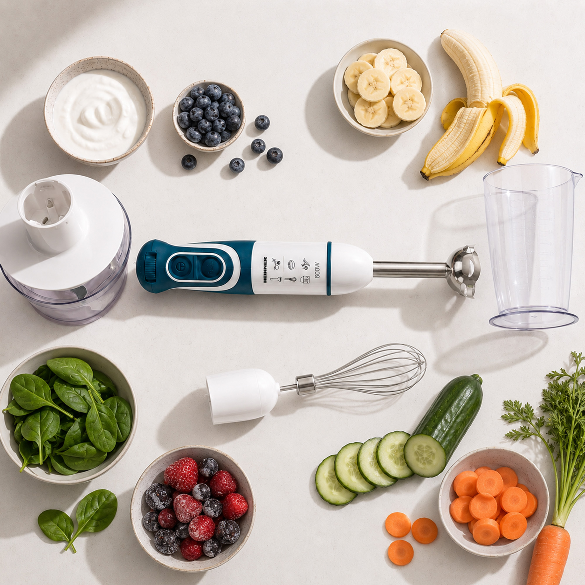 Ein Stabmixer mit Zubehör, Joghurt, Beeren, Bananen, Spinat, Gurken und Karotten.