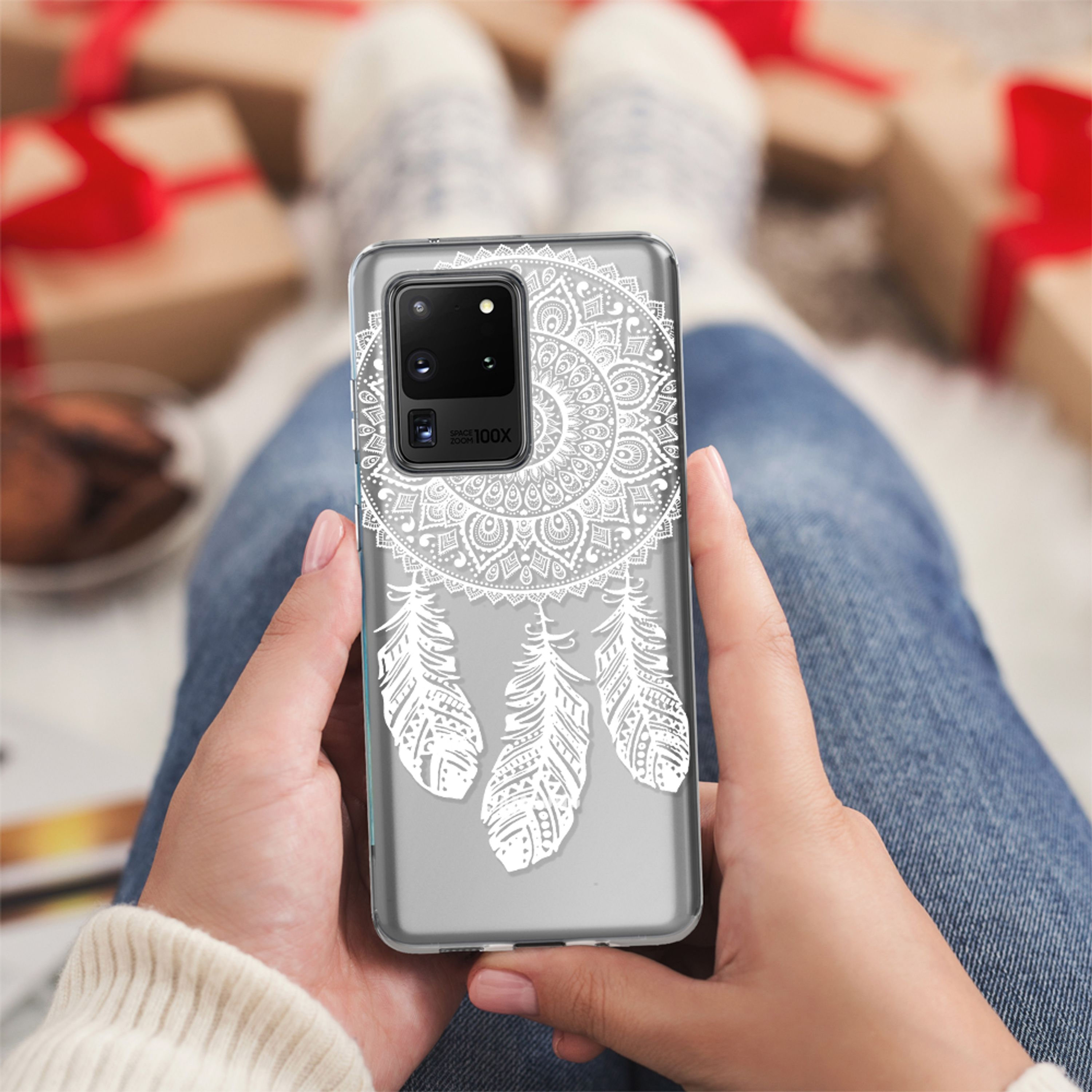 Person hält ein Handy mit weißem Federmuster. Geschenke im Hintergrund.