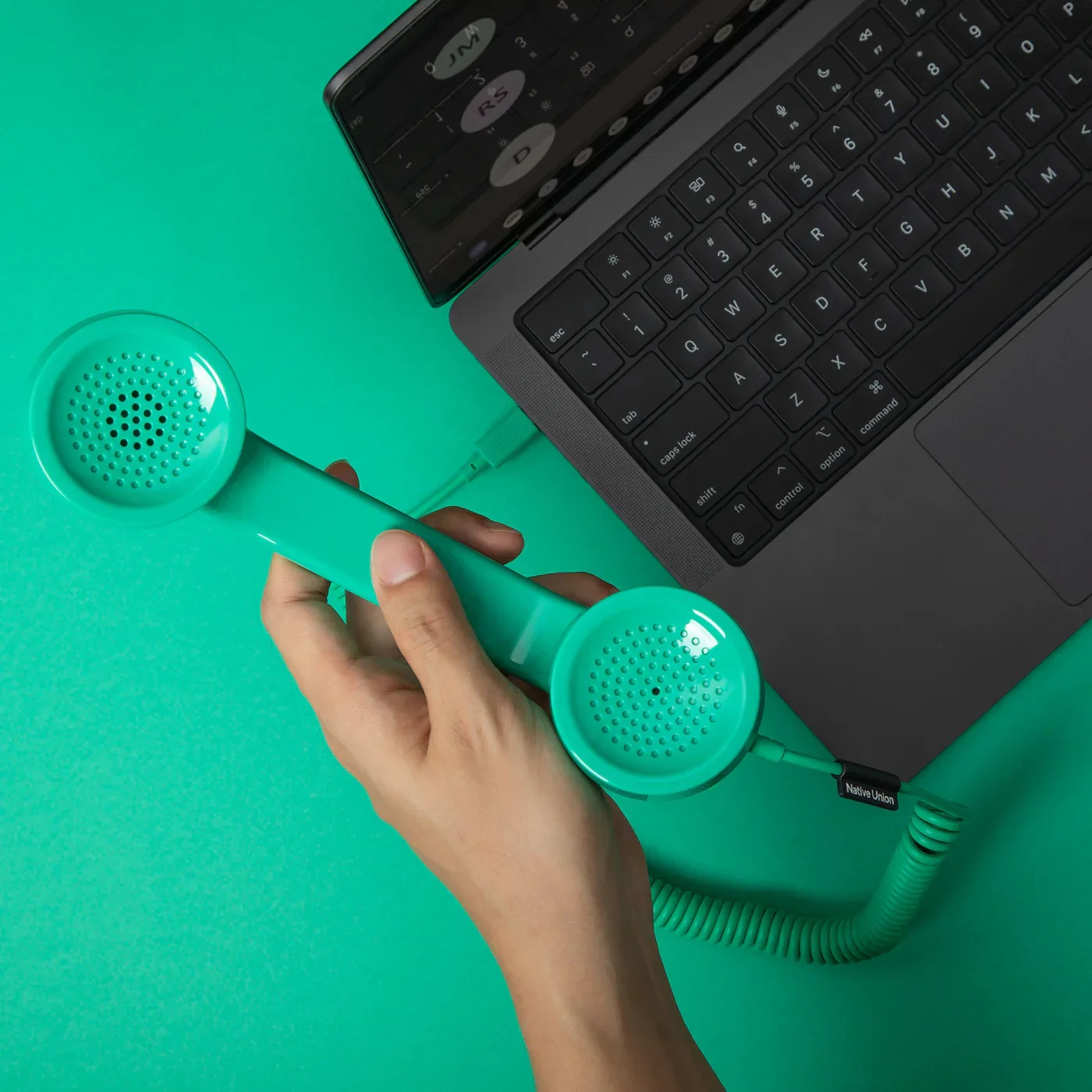 Una mano tiene una cornetta telefonica verde acqua accanto a un laptop aperto su sfondo verde acqua.