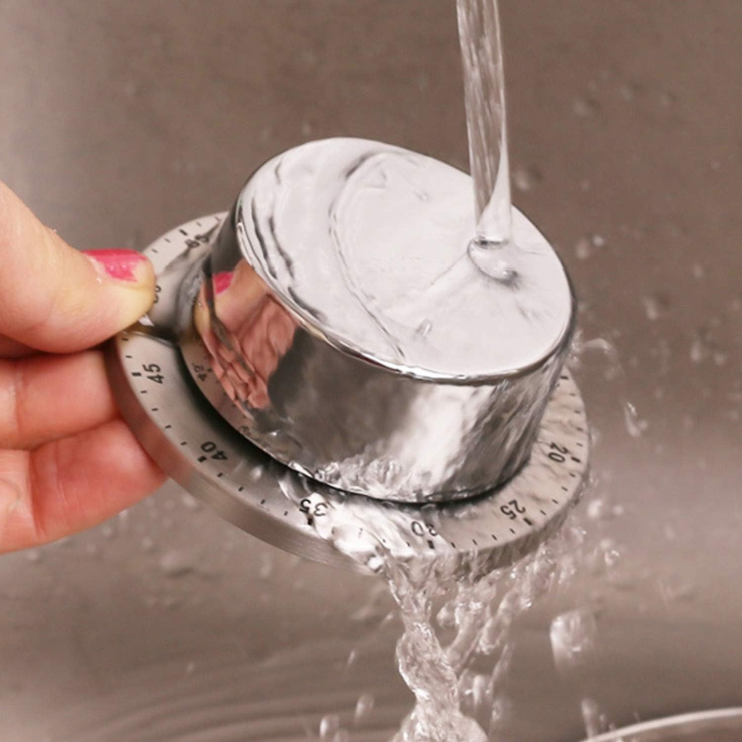 Eine Hand mit rosa Nagellack dreht eine silberne Küchenuhr unter einem Wasserstrahl.