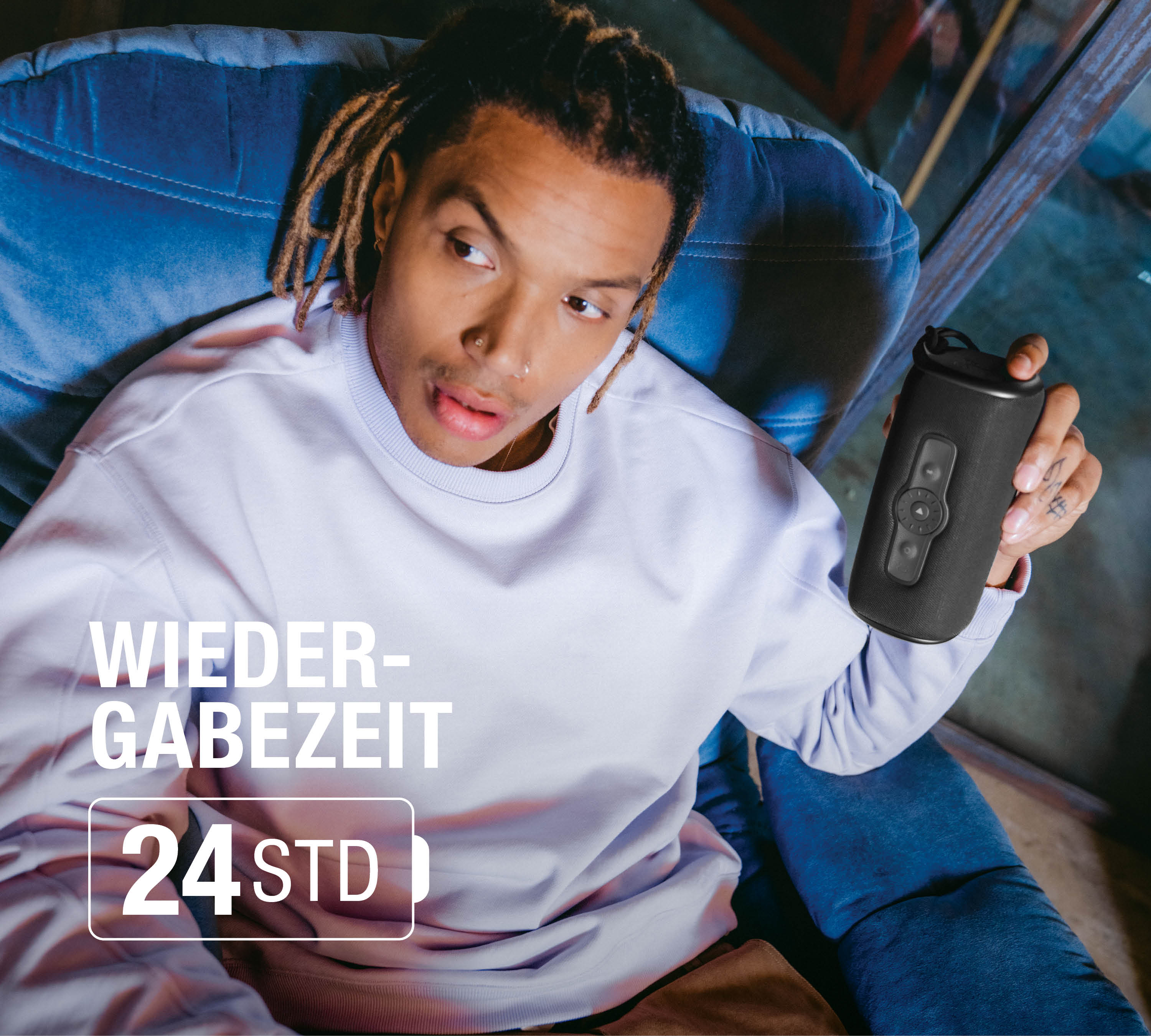 Mann mit Dreadlocks hält schwarzen Lautsprecher in blauem Sessel. Text: 24 Std.