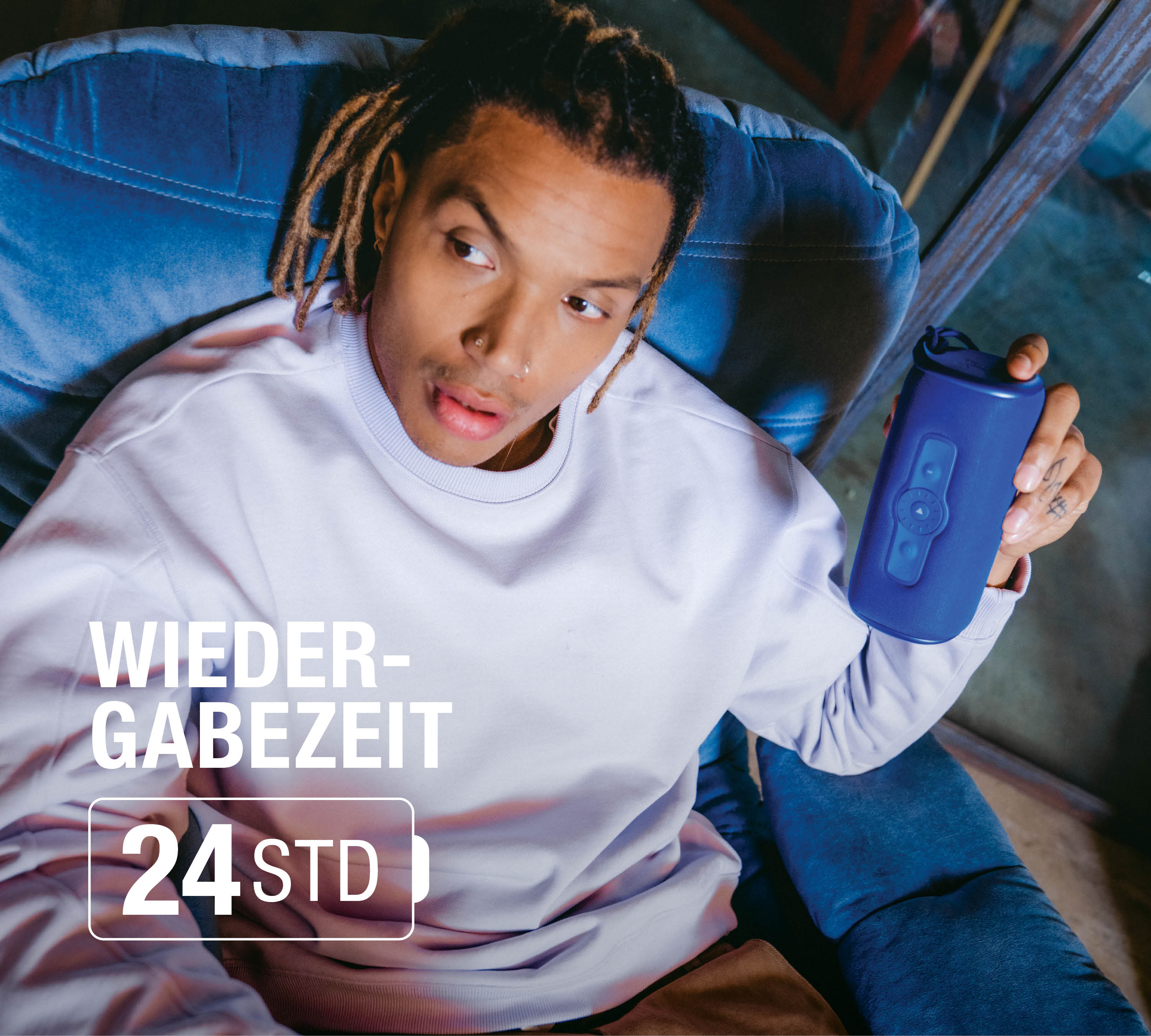 Mann mit Dreadlocks sitzt im Sessel und hält einen blauen Lautsprecher.