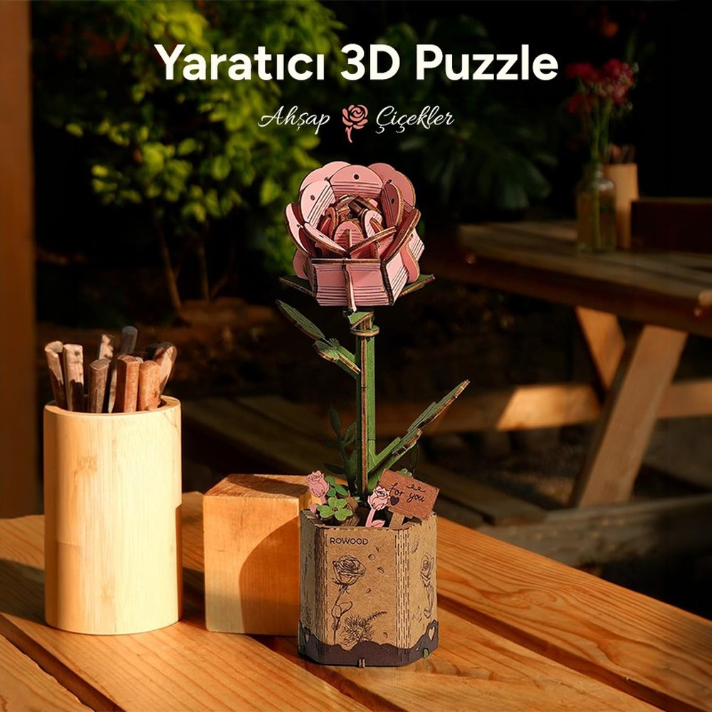 Un puzzle de rosa 3D rosa está en una maceta sobre una mesa de madera. Al lado, un recipiente de madera con palitos.