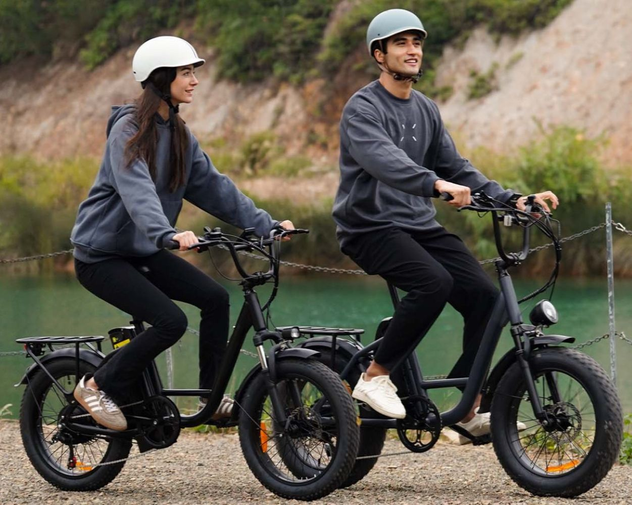Hombre y mujer montan bicicletas eléctricas junto a un lago, ambos con casco.