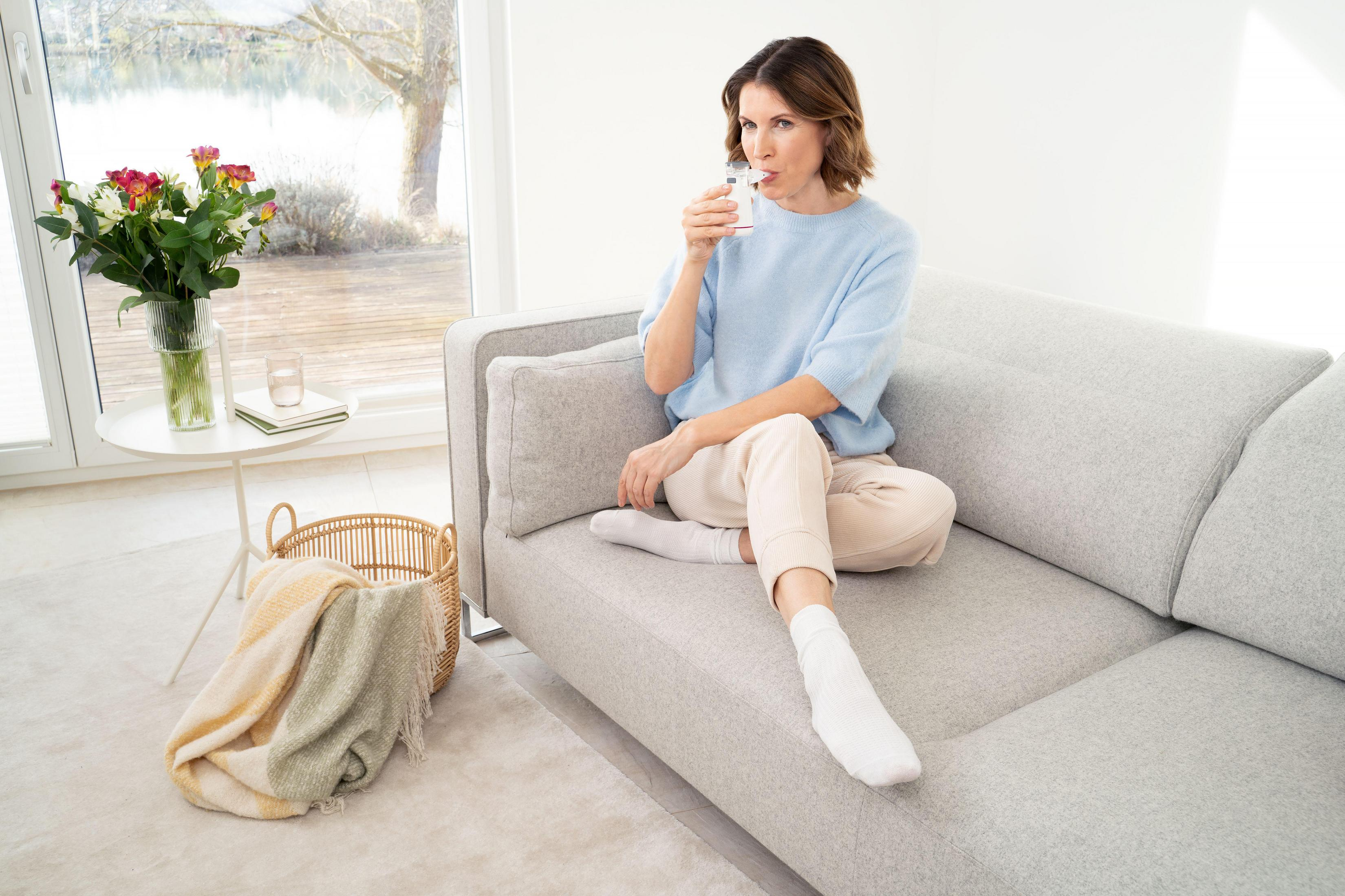 Eine Frau sitzt auf einem Sofa und trinkt aus einem Glas. Blumen stehen auf einem Tisch.