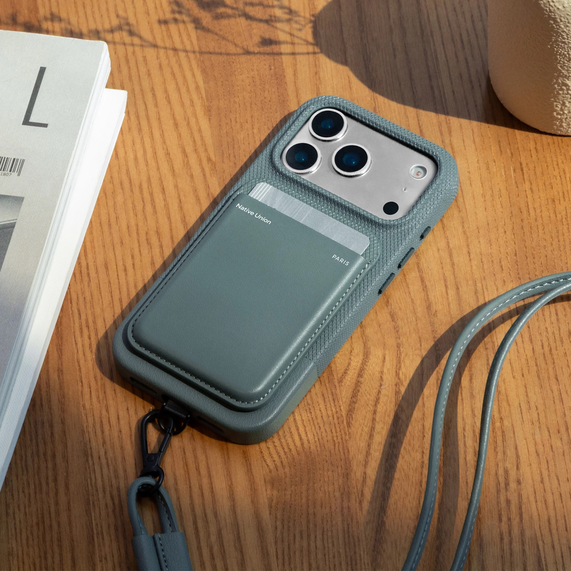 Un telefono grigio con porta carte e cordino poggia su un tavolo di legno accanto a libri.