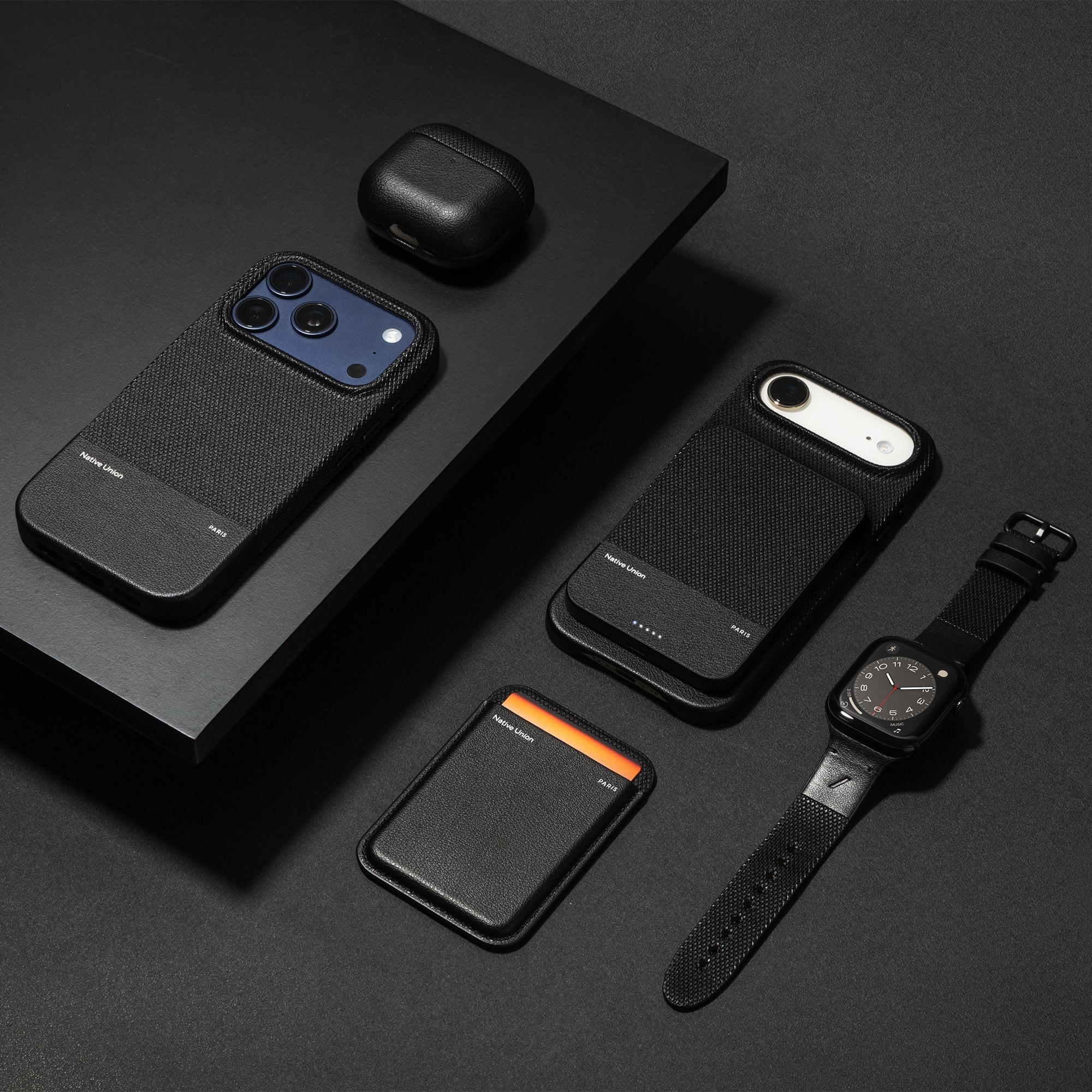 Schwarze Handyhülle, Kopfhörerhülle, Kartenetui und Smartwatch auf schwarzem Hintergrund.