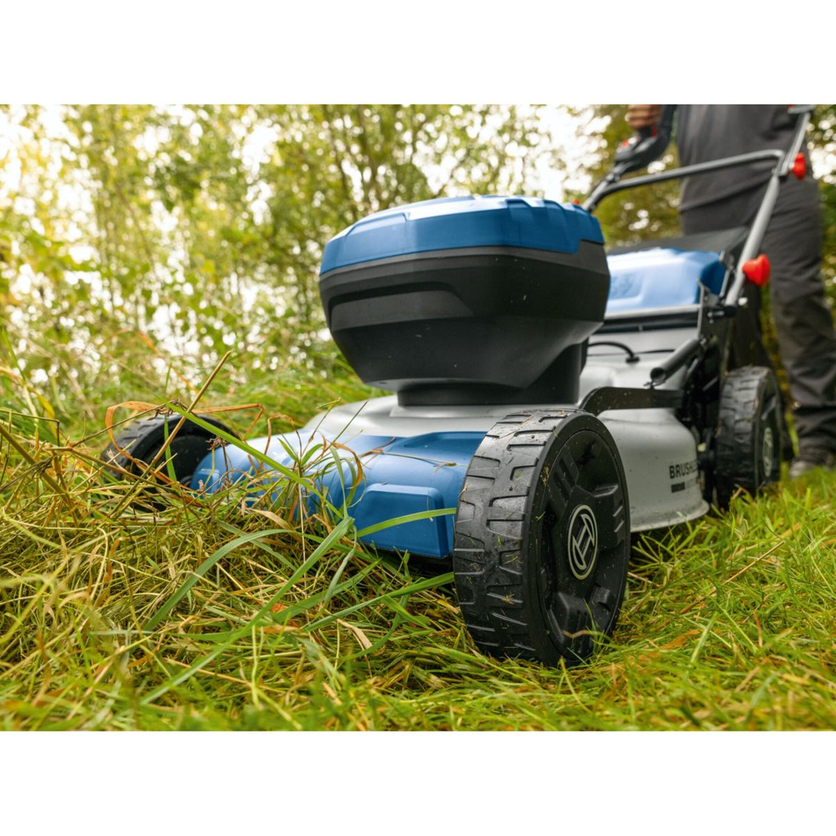 Ein blauer und grauer Rasenmäher wird von einer Person durch hohes Gras geschoben.