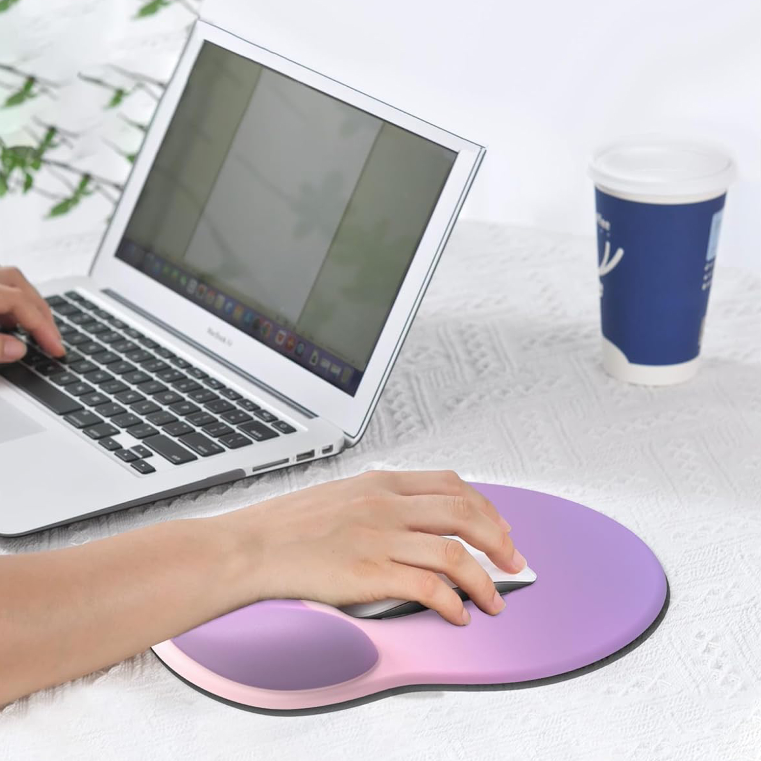 Person tippt auf einem Laptop neben einer blauen Kaffeetasse. Ein lila Mauspad ist im Vordergrund.