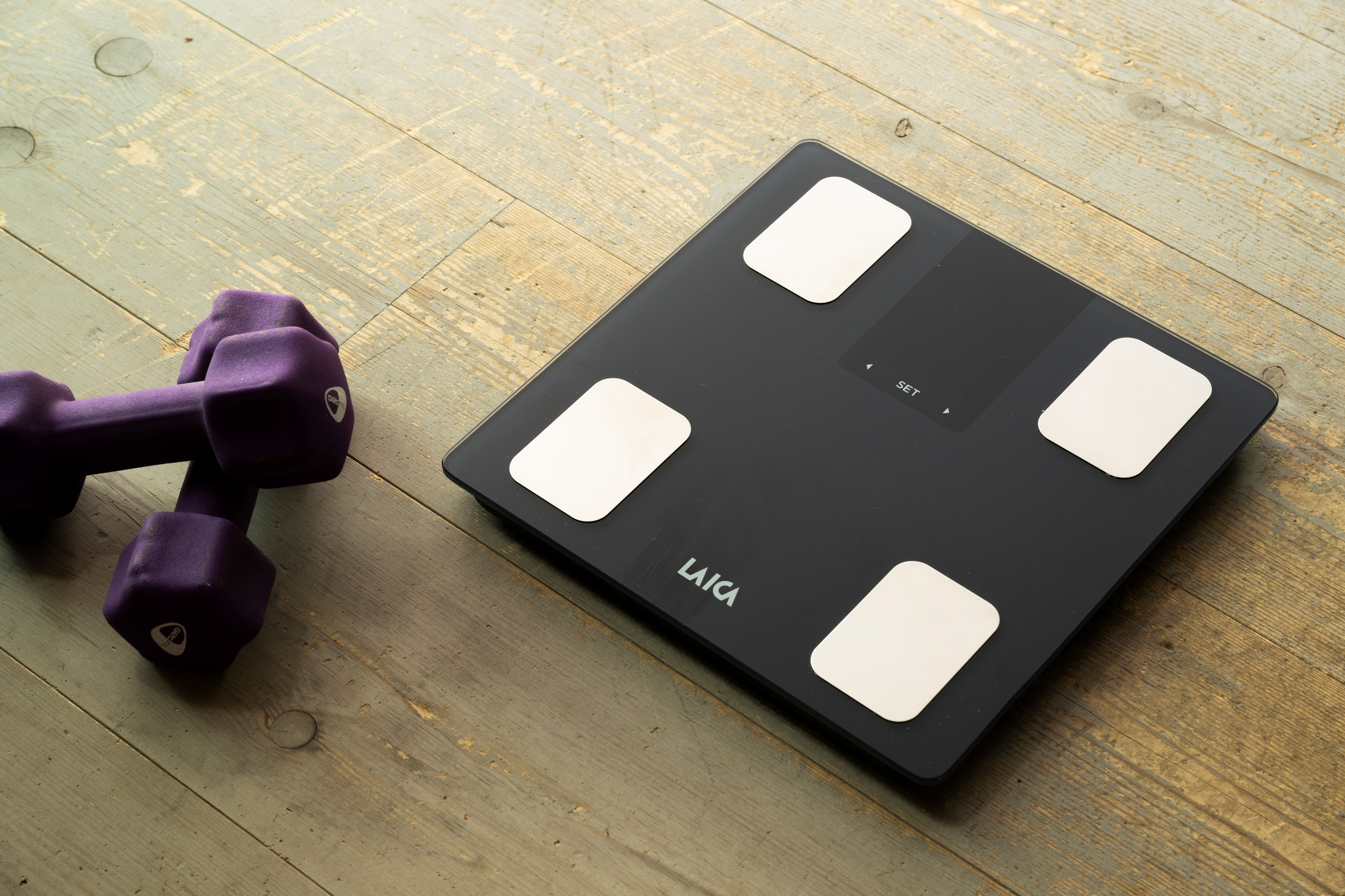 Báscula digital negra y mancuernas moradas en suelo de madera.