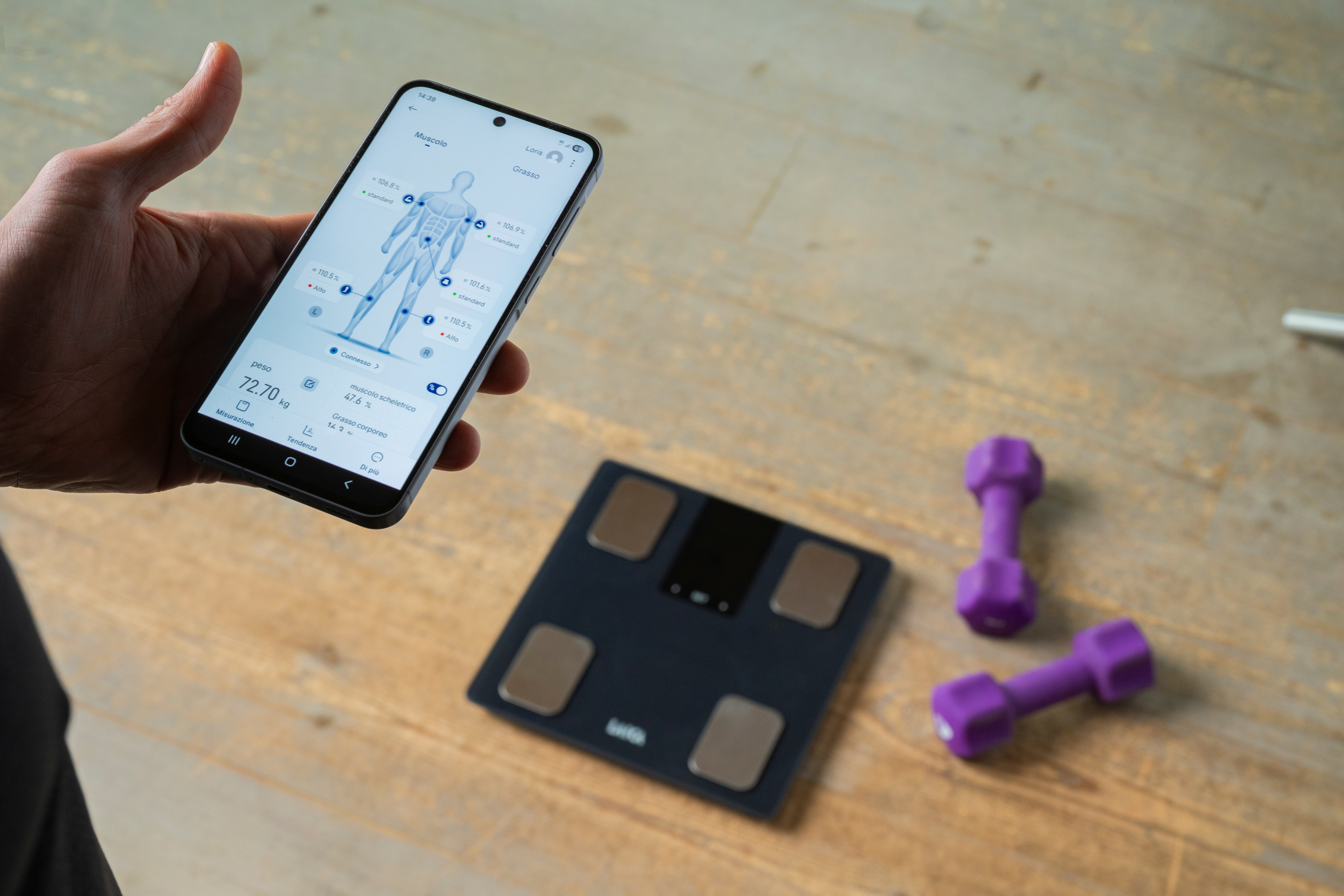 Una mano sostiene un smartphone que muestra datos corporales. Una báscula y mancuernas están cerca.