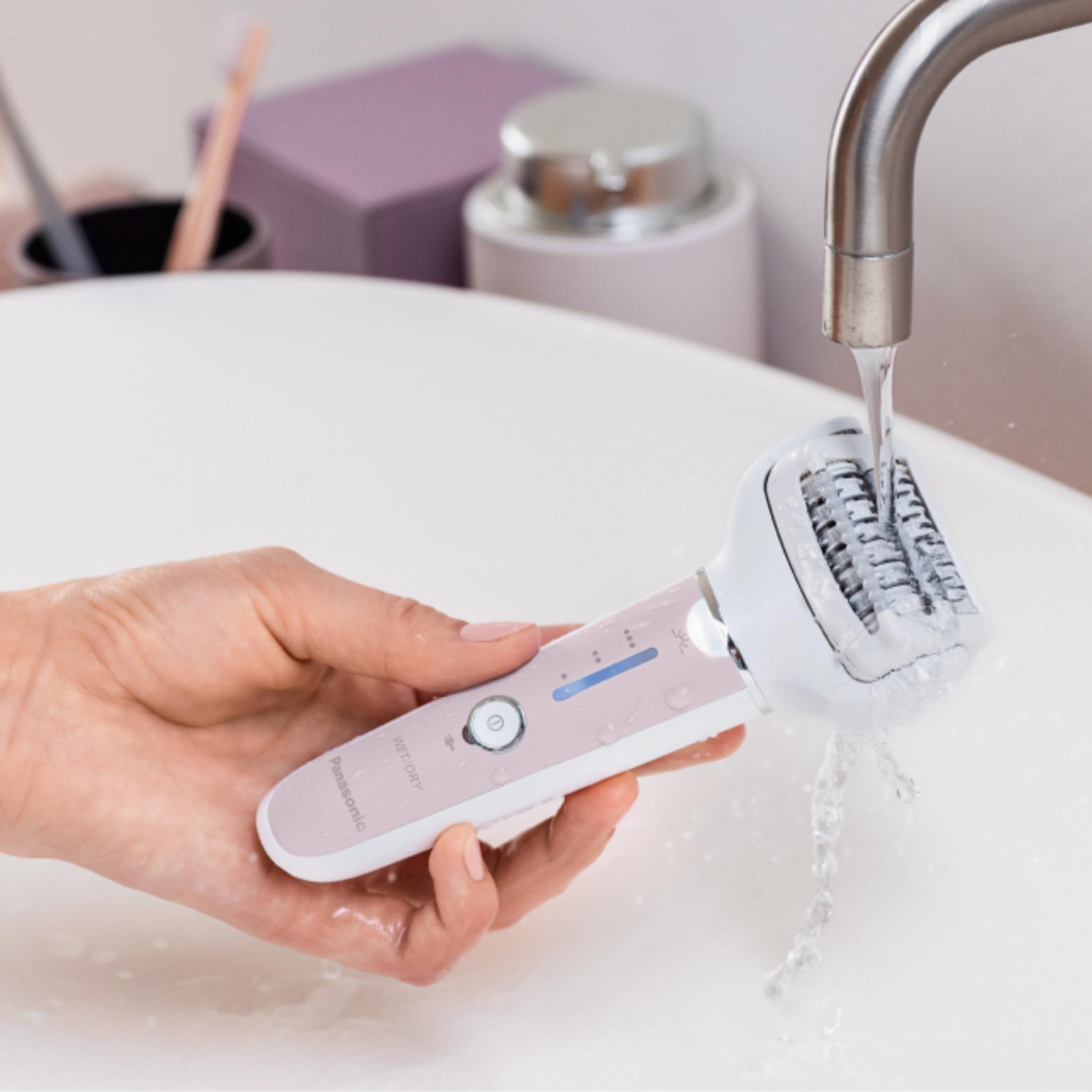 Una mano tiene un epilatore rosa sotto acqua corrente in un lavandino.