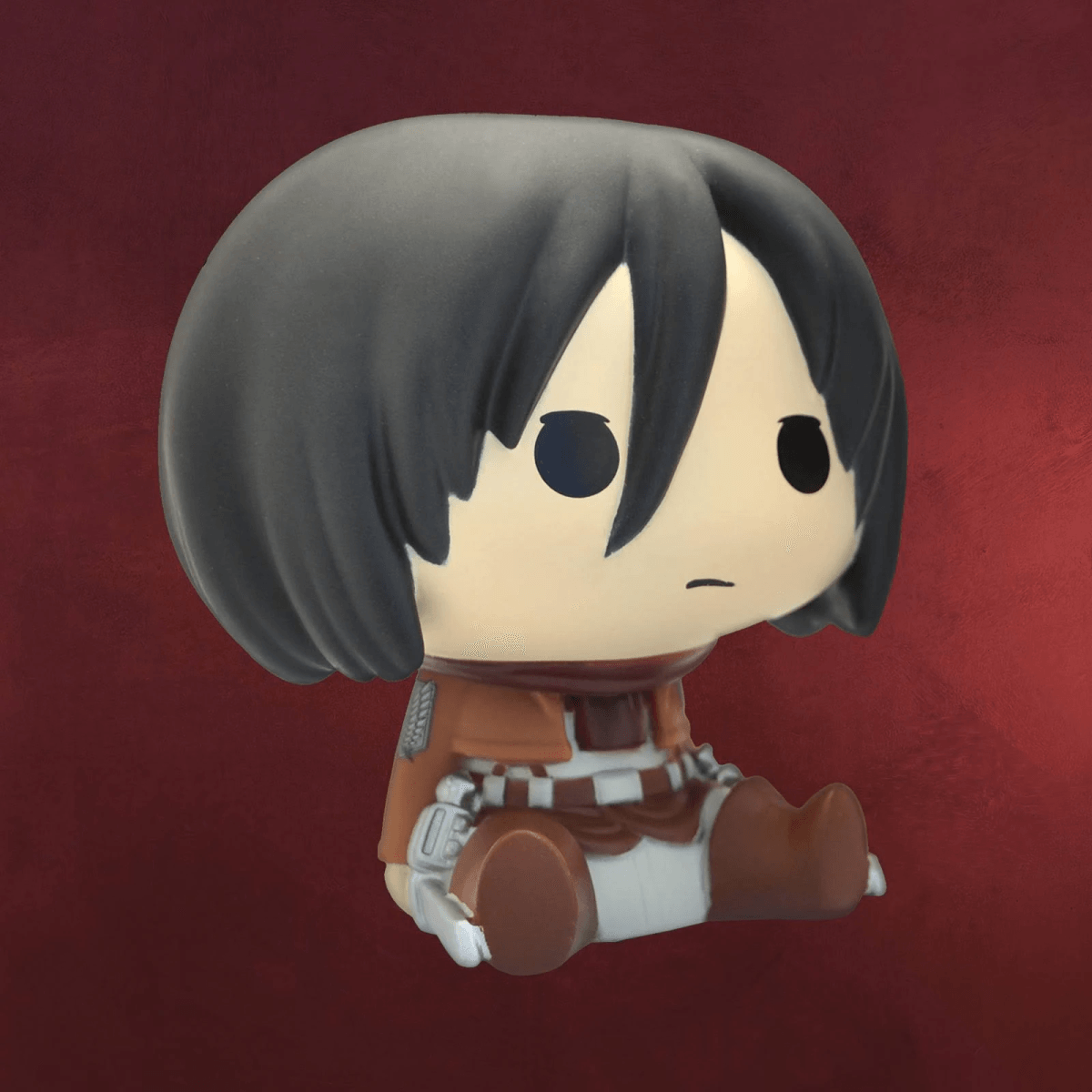 Figura chibi de Mikasa con capa y uniforme marrones, sentada sobre fondo rojo.