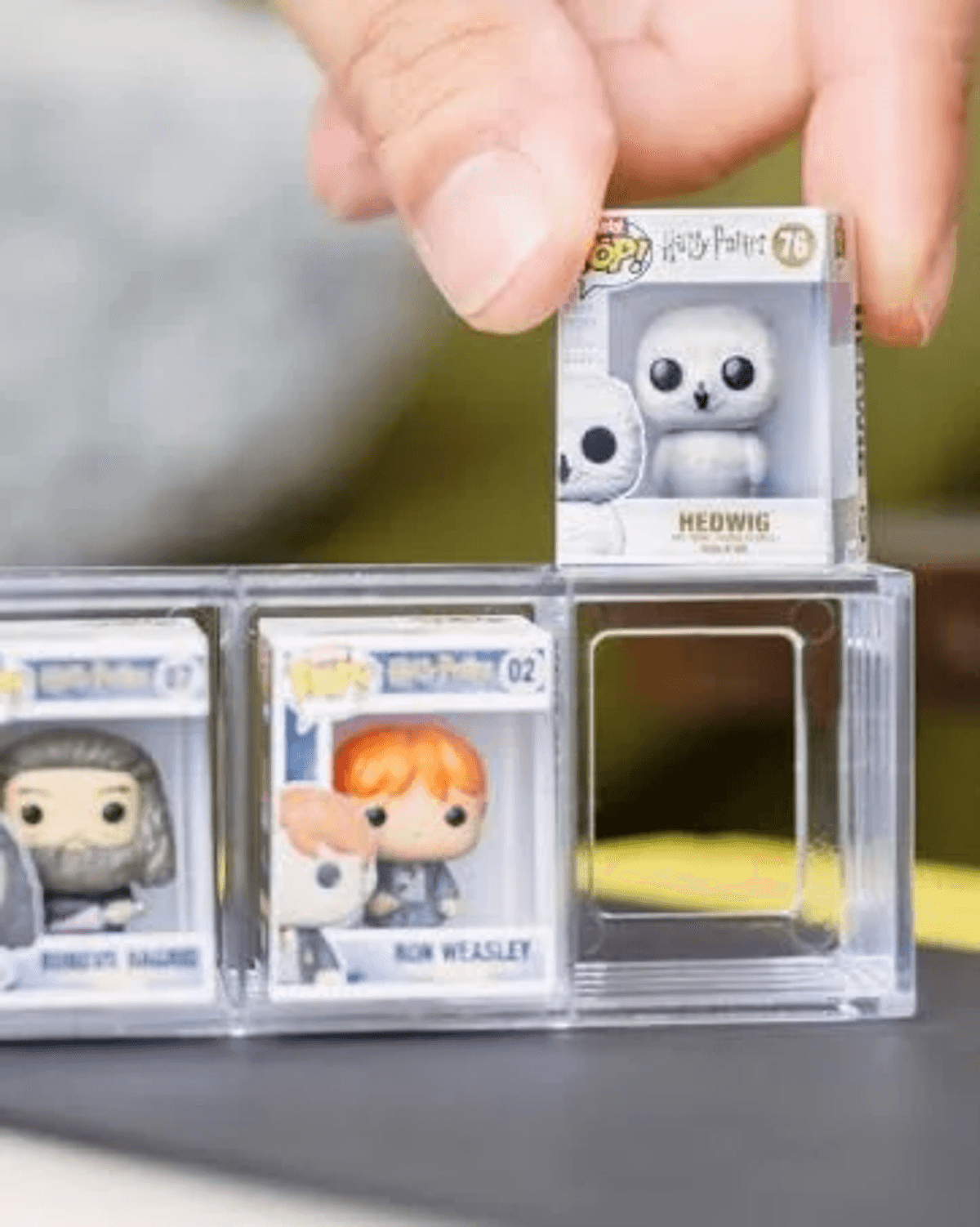 Una mano coloca un Funko Pop blanco de Hedwig en una vitrina transparente, junto a Ron Weasley y Hagrid.