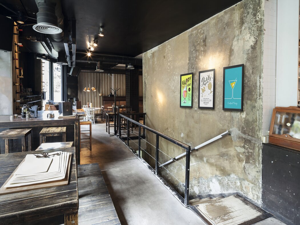 Ein Restaurant mit rustikalen Holztischen und Barhockern, Betonwänden und Cocktail-Postern.
