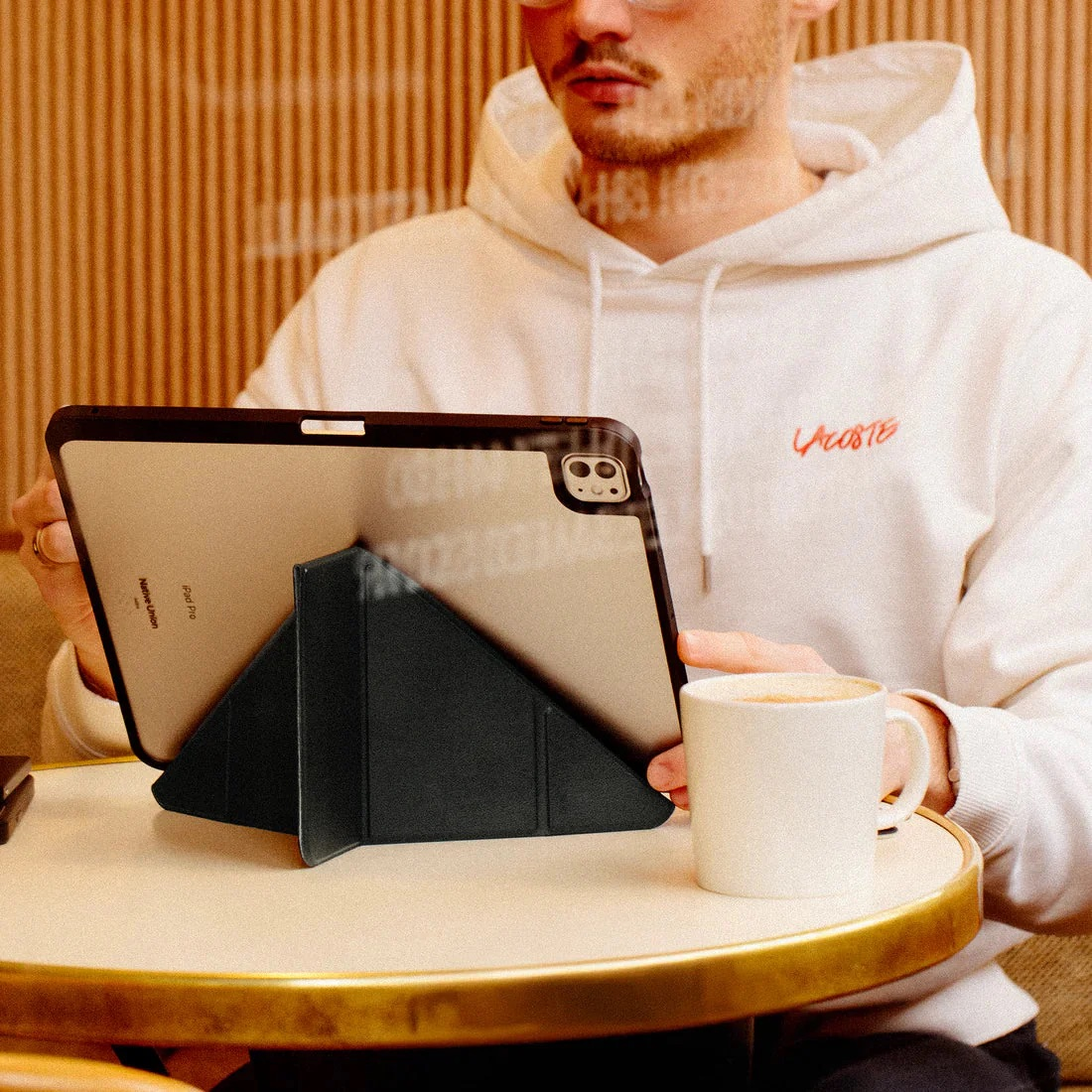 Mężczyzna w bluzie z kapturem trzyma tablet na stojaku obok filiżanki kawy.