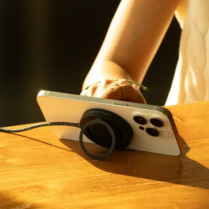 Un iPhone blanco con soporte de anillo negro, sobre mesa de madera. Mano presente.