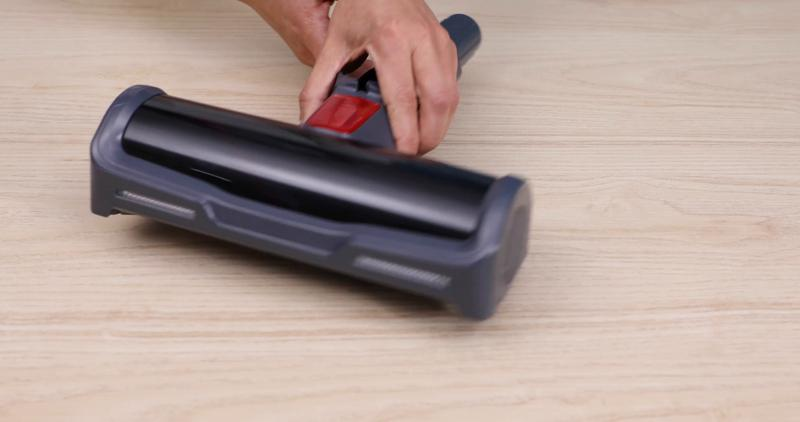 Una mano sostiene un accesorio de aspiradora oscuro con palanca roja sobre suelo de madera.