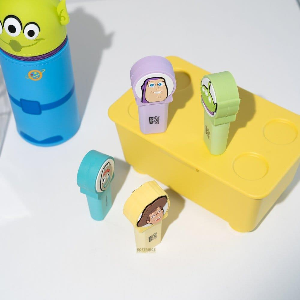 Toy Story Figuren auf gelber Kiste und Alien-Stifte auf weißem Tisch.
