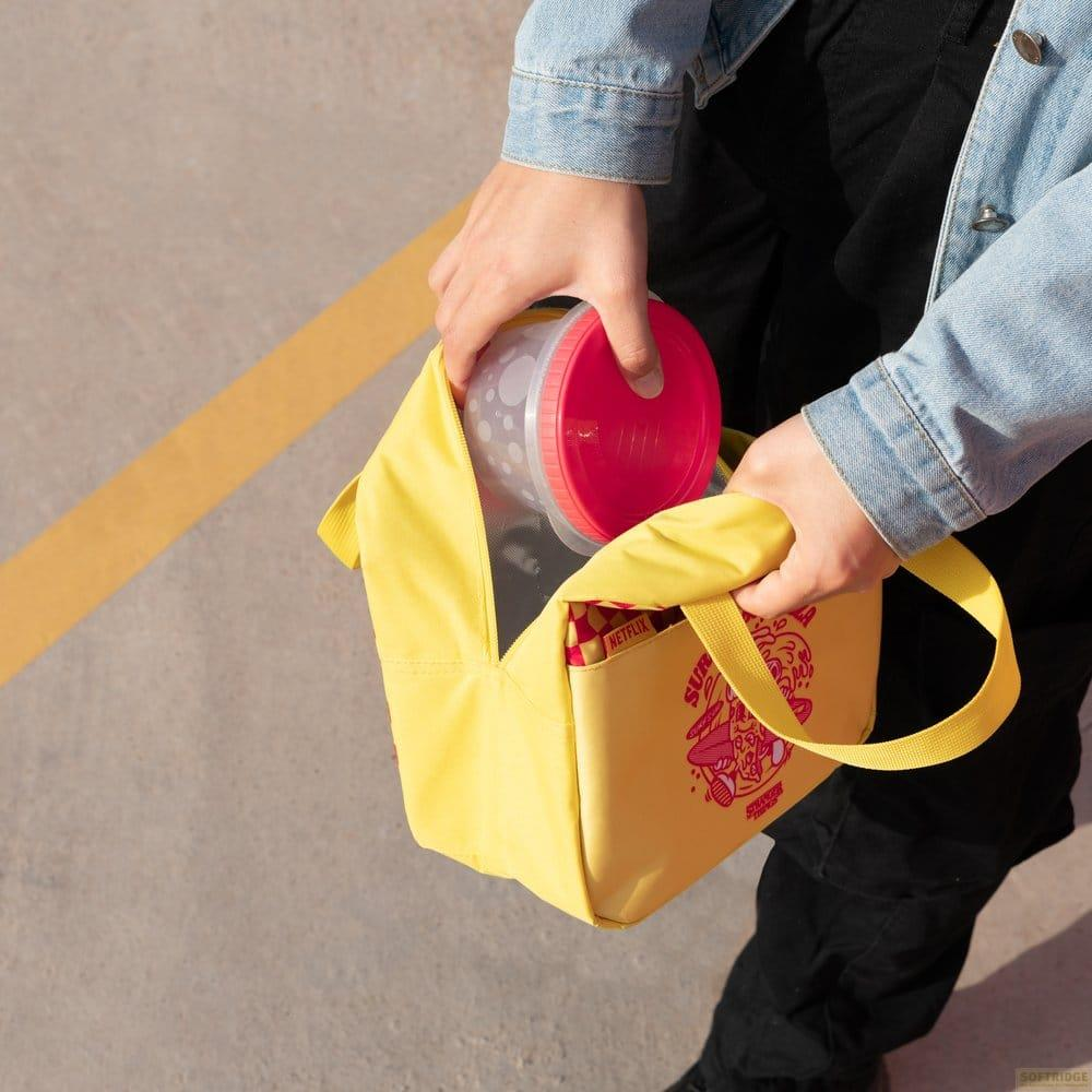 Person packt einen Lebensmittelbehälter mit rotem Deckel in eine gelbe Stranger Things Lunchtasche.