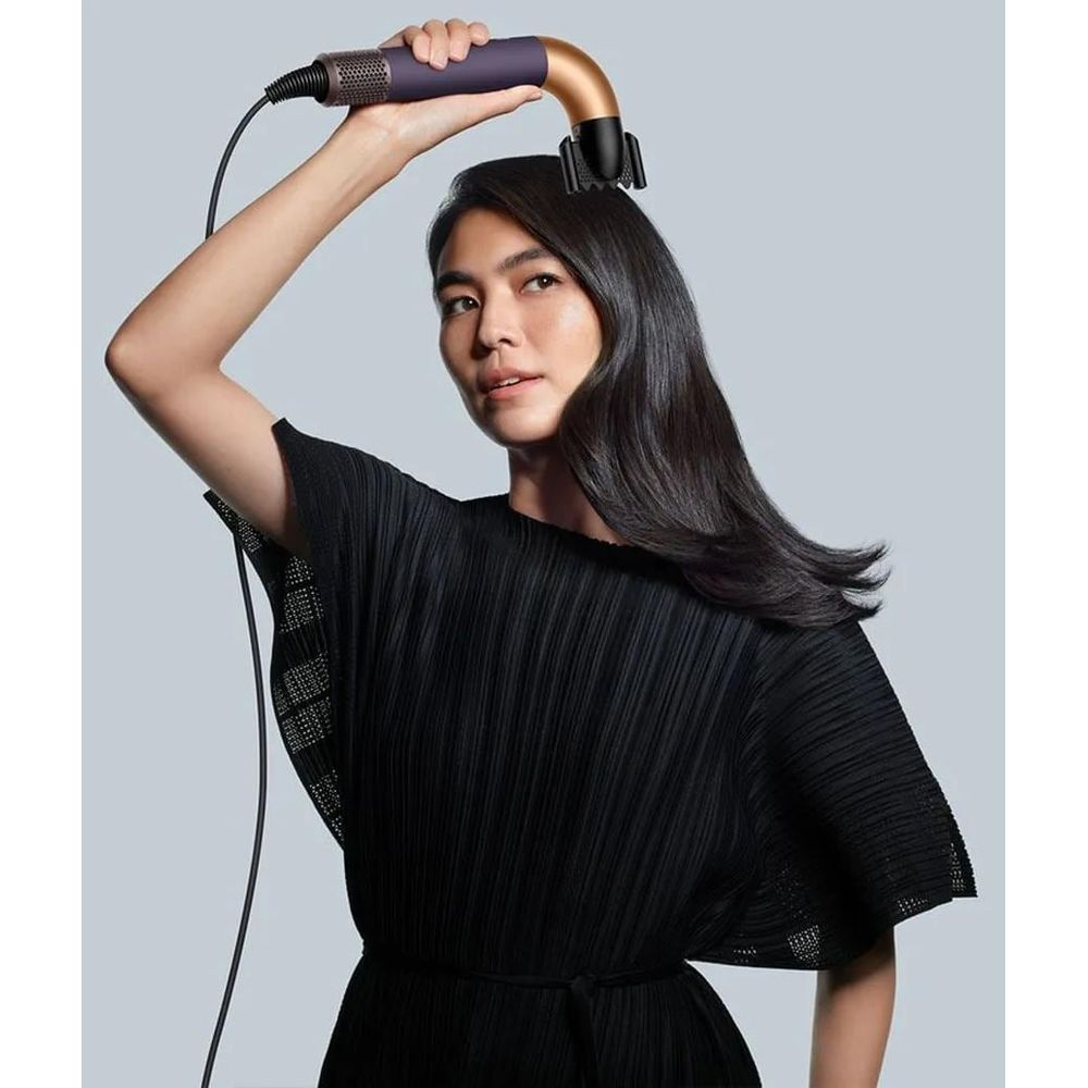 Donna in abito nero usa un asciugacapelli viola e oro sui suoi lunghi capelli scuri.