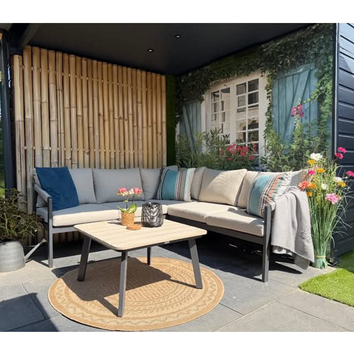 Een L-vormige loungebank met tafel staat op een terras naast een bamboescherm en een huis.