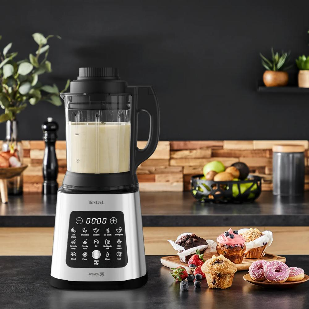 Ein silberner Tefal-Mixer mit vollem Krug steht auf einer Theke, mit Muffins, Donuts und Obst.