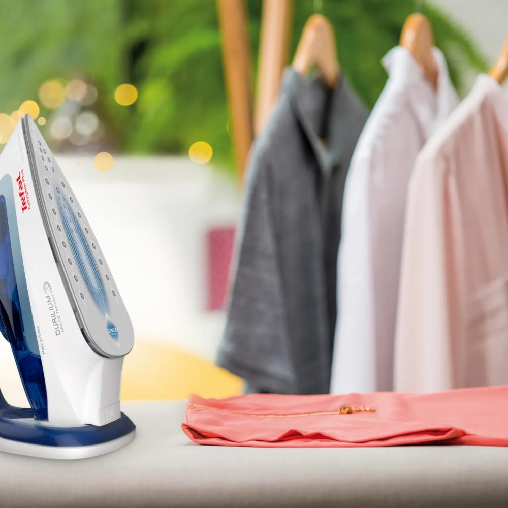 Ein Tefal-Bügeleisen ruht auf einem Tisch neben gefaltener Korallen-Stoff. Kleidung hängt verschwommen im Hintergrund.