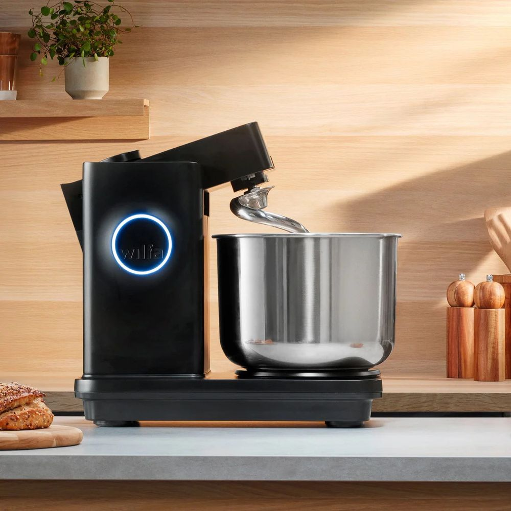 Impastatrice nera con ciotola in acciaio, logo illuminato blu, sfondo in legno.