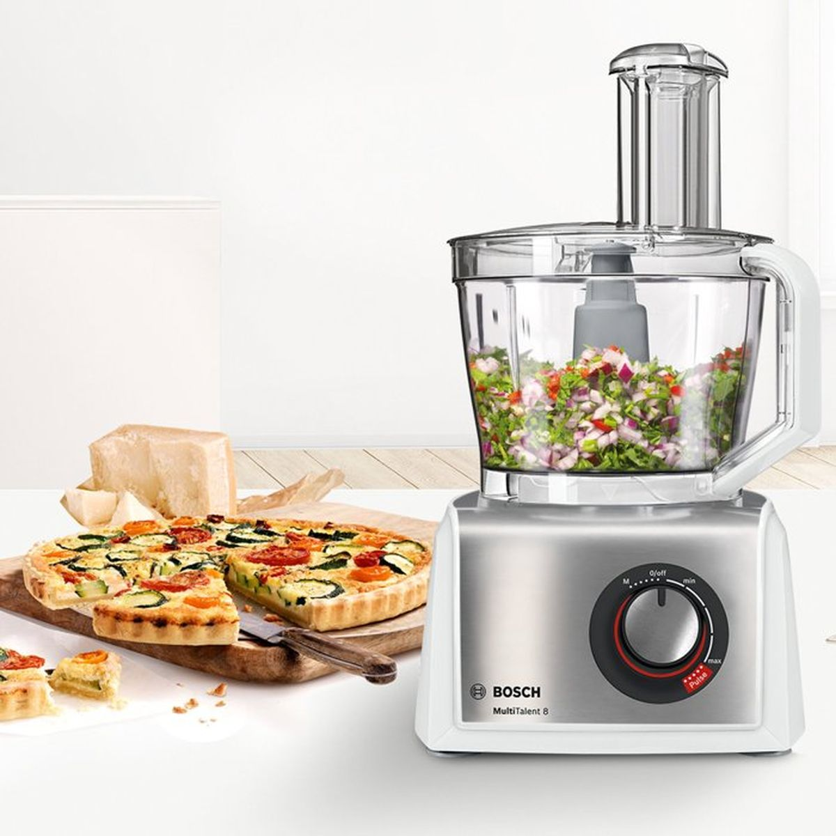 Un robot da cucina con verdure tritate, accanto a una quiche e pezzi di formaggio.