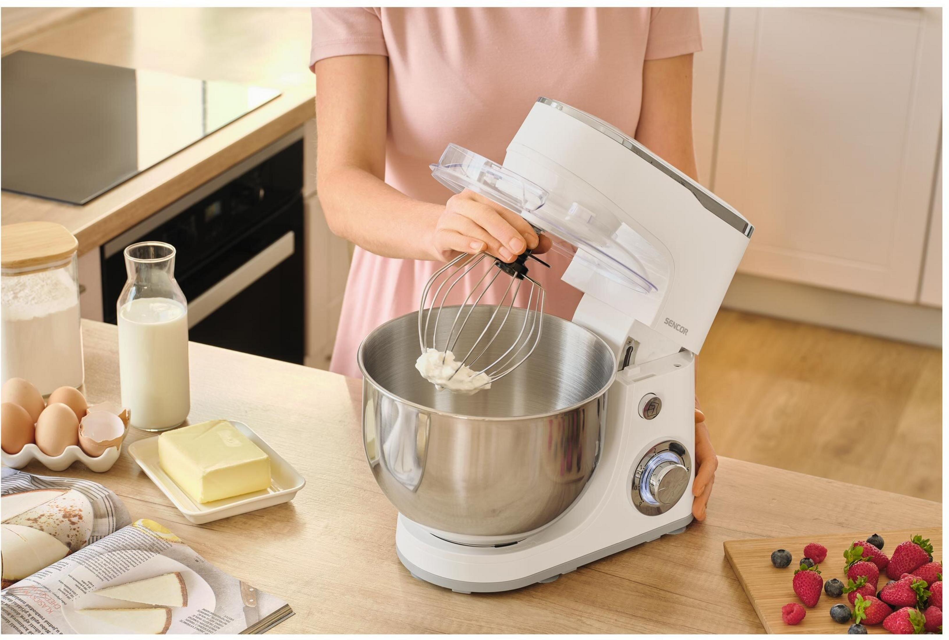 Una donna usa un'impastatrice planetaria bianca con frusta. Ingredienti come uova, latte e burro sono pronti.