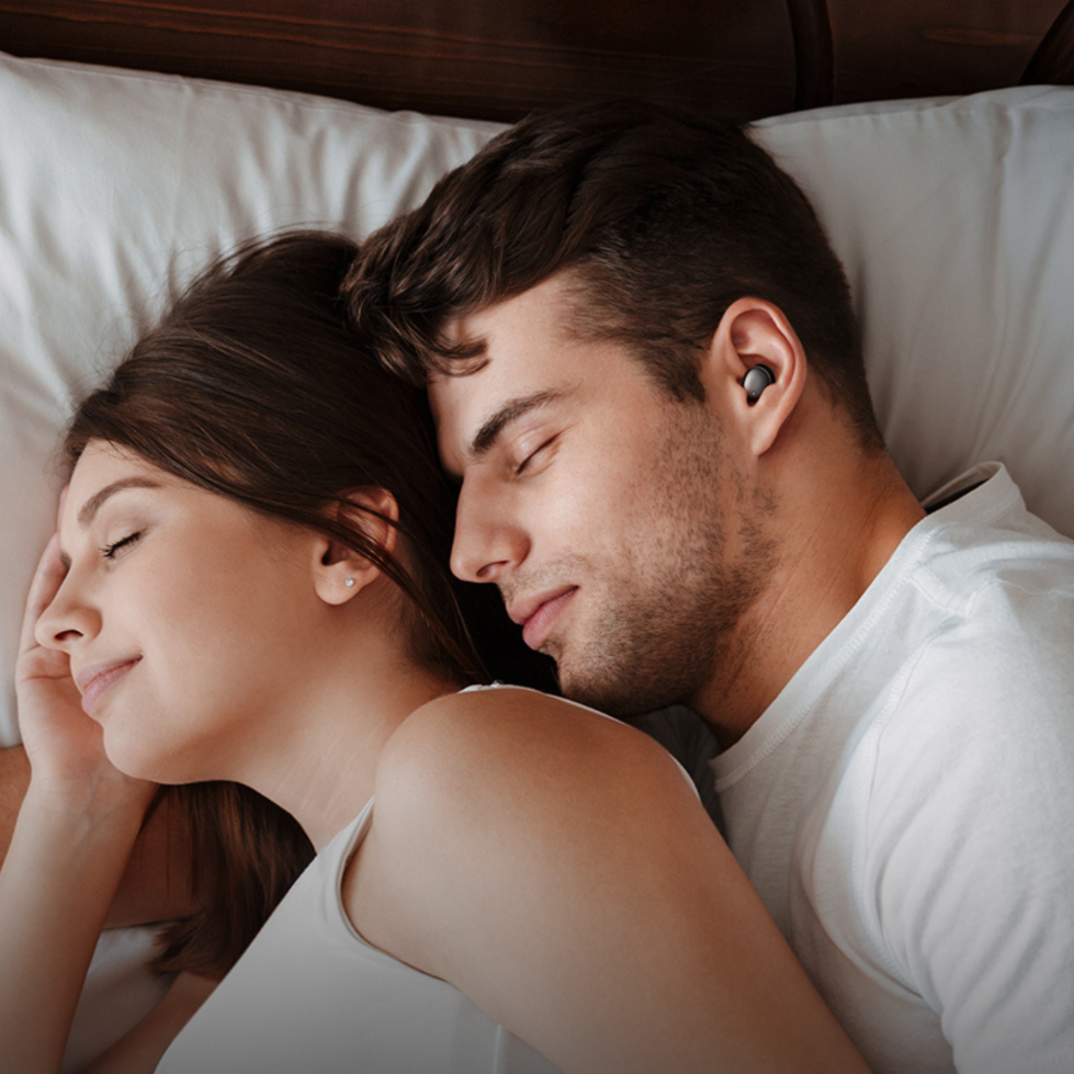 Coppia dorme a letto, uomo indossa auricolari neri. Donna appoggia testa sulla sua spalla.