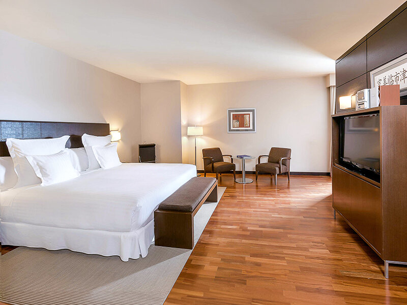 Una cama grande y hecha con sábanas y almohadas blancas está en primer plano de una habitación de hotel.