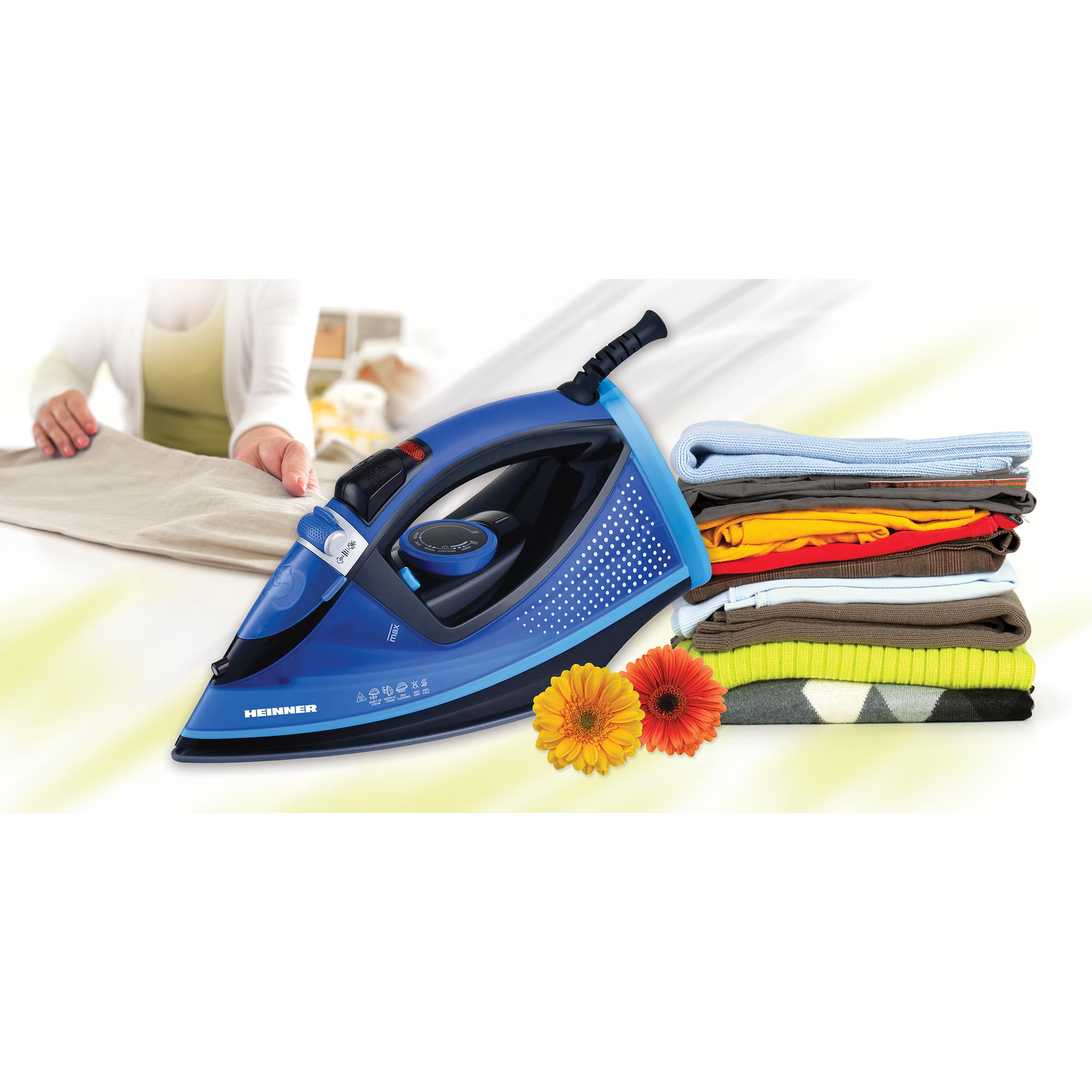 A blue Heinner iron sits beside a stack of colorful clothes and two flowers.