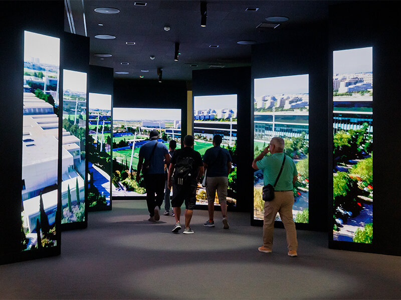 Gente camina por una exposición con pantallas verticales que muestran un paisaje urbano.