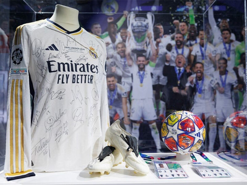 Camiseta de fútbol firmada, botines y balón en un museo con jugadores vitoreando al fondo.
